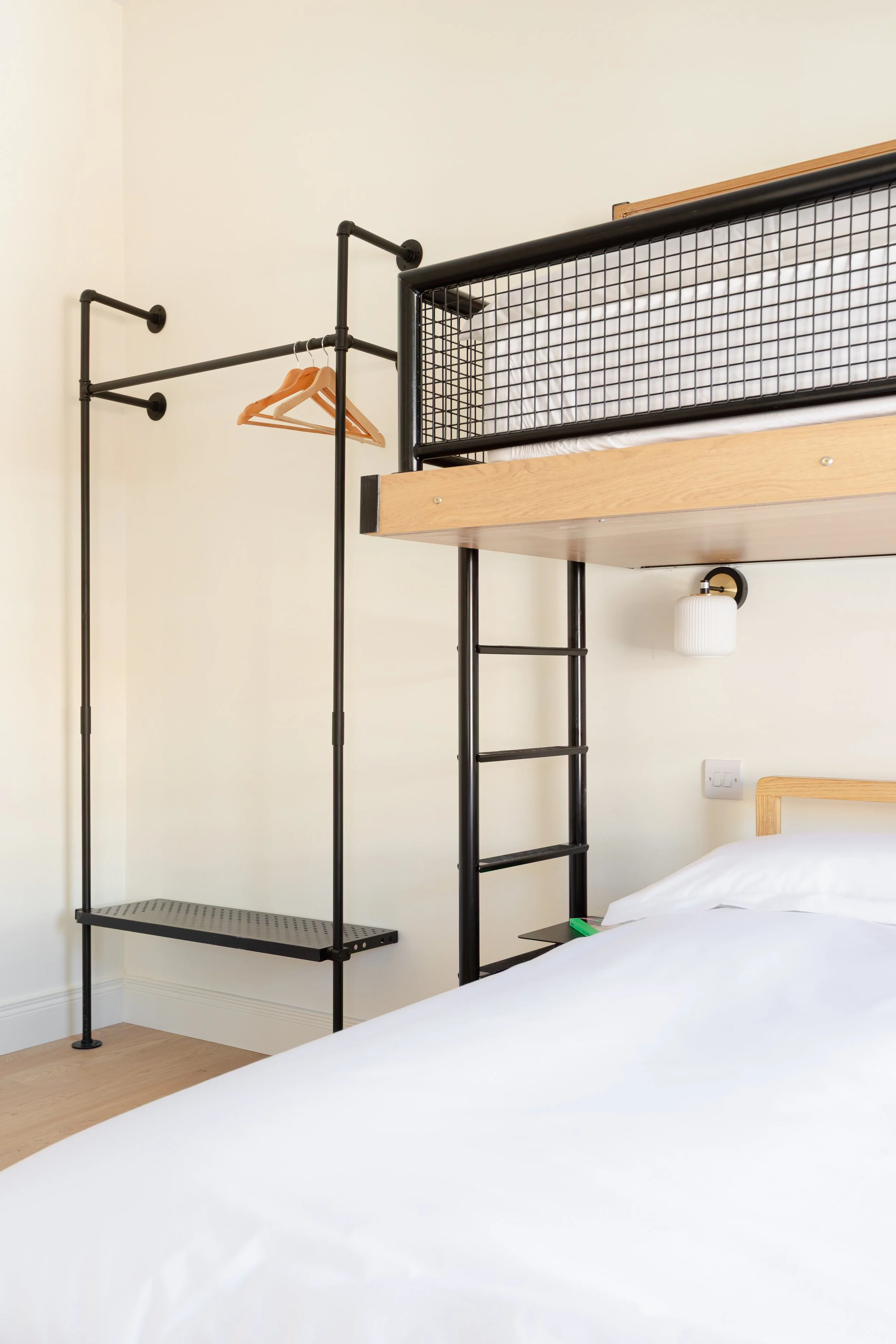 A bedroom with a loft bed featuring black metal and wood construction. The bed has a white mattress and a black wire guardrail. There is a ladder on the side of the bed. Next to the bed is a wall-mounted lantern-style light fixture. An open closet with a black metal clothing rack, wooden hangers, and a perforated black metal shelf is visible in the corner.