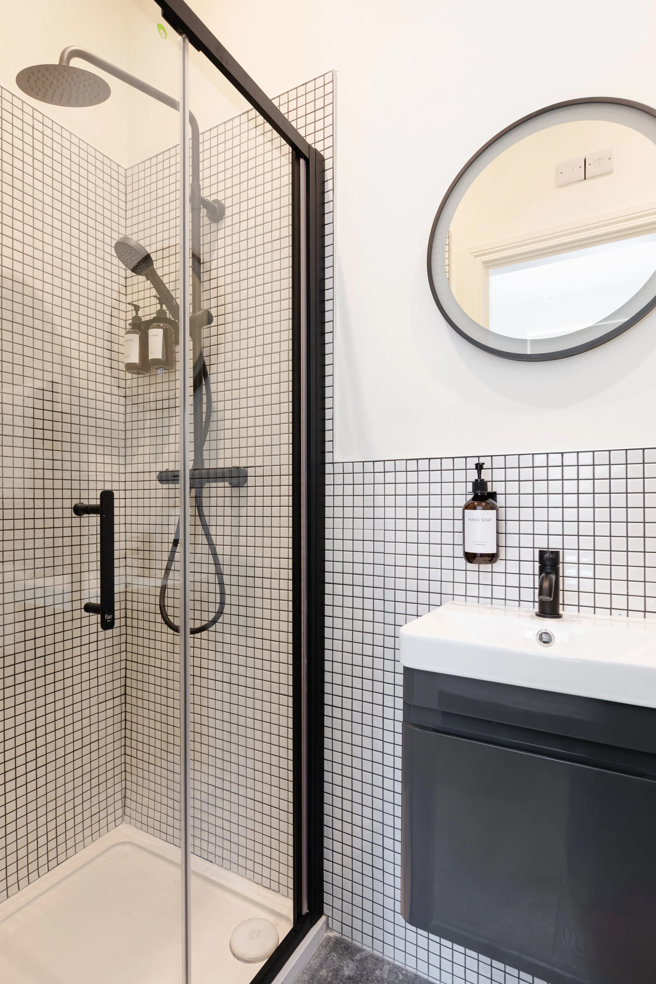 Modern bathroom with a corner shower featuring small black and white square tiles, a round mirror, a dark grey vanity with a white sink, a black faucet, and a hand soap dispenser.