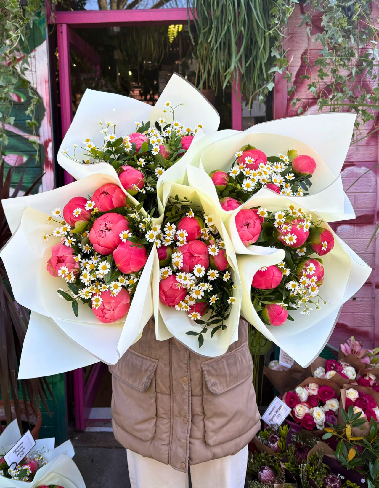 Person holding a large bouquet of pink peonies and white daisies in a flower shop.