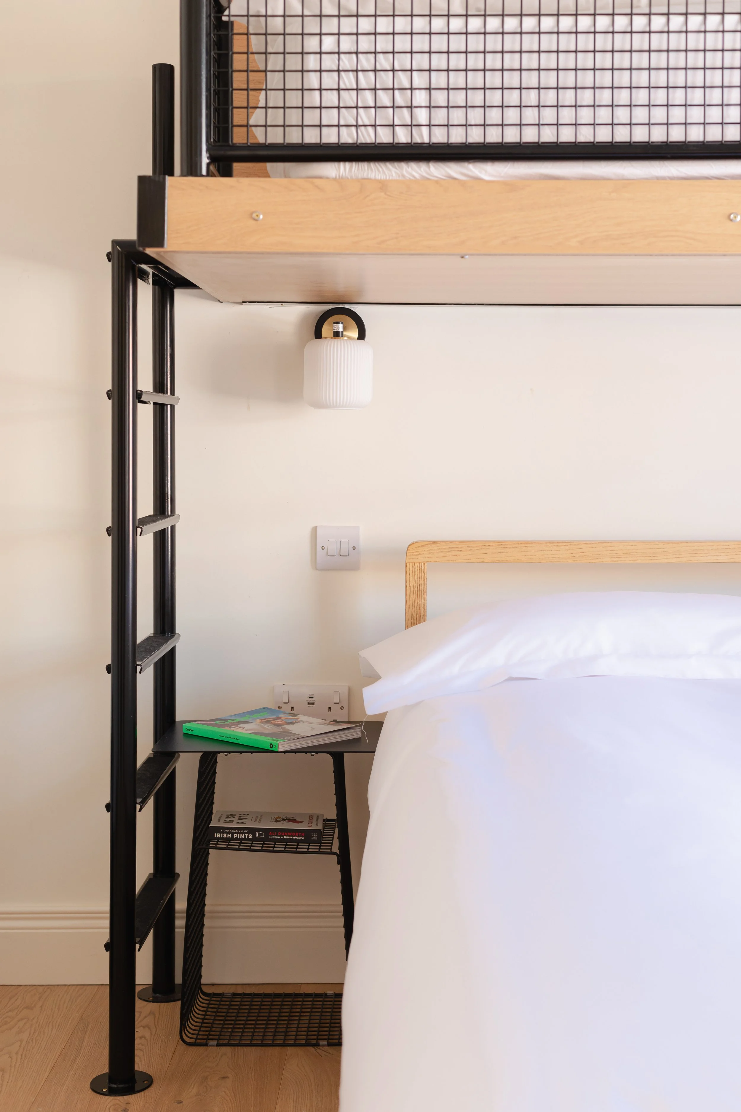 A bedroom corner with a black metal and wood loft bed, a nightstand with magazines, and a wall-mounted light fixture.