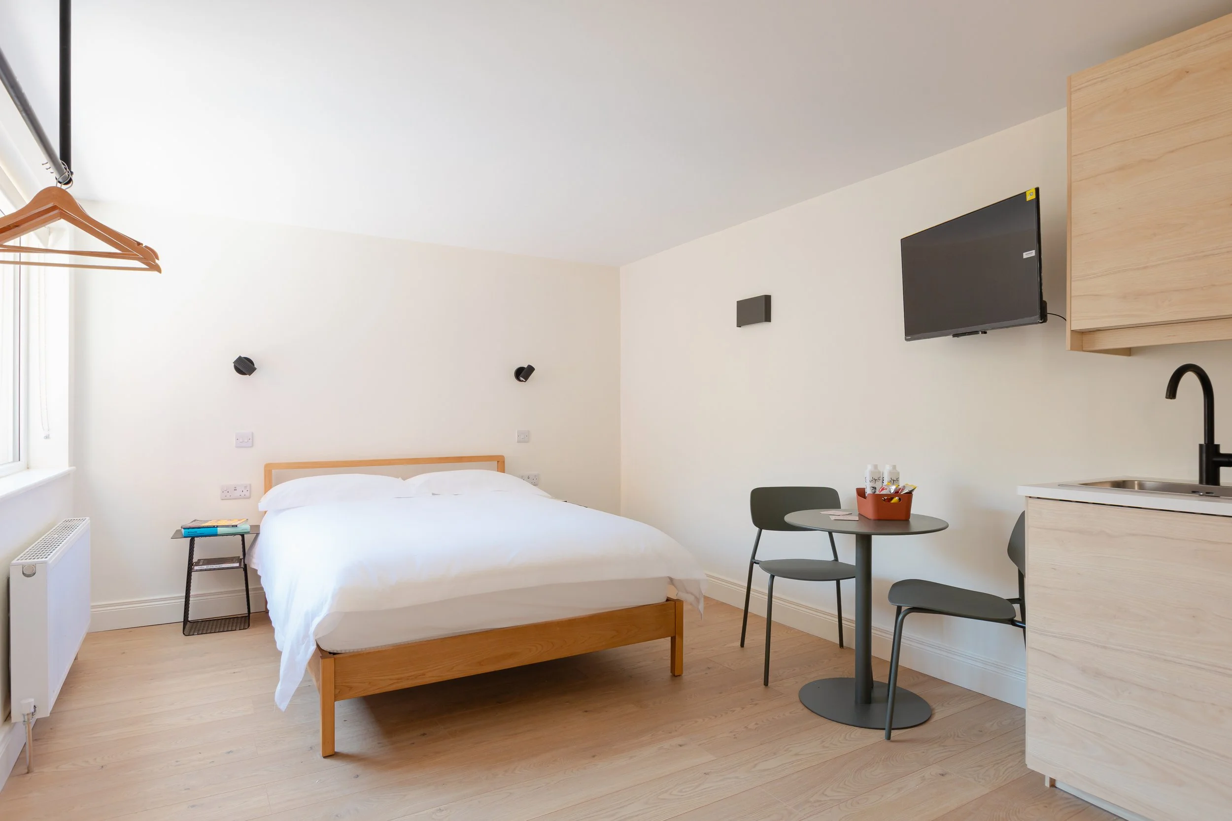 A bedroom with a wooden bed frame, white bedding, a small side table with books, a wall-mounted TV, a small table with two chairs, and a partial kitchenette area with a sink.