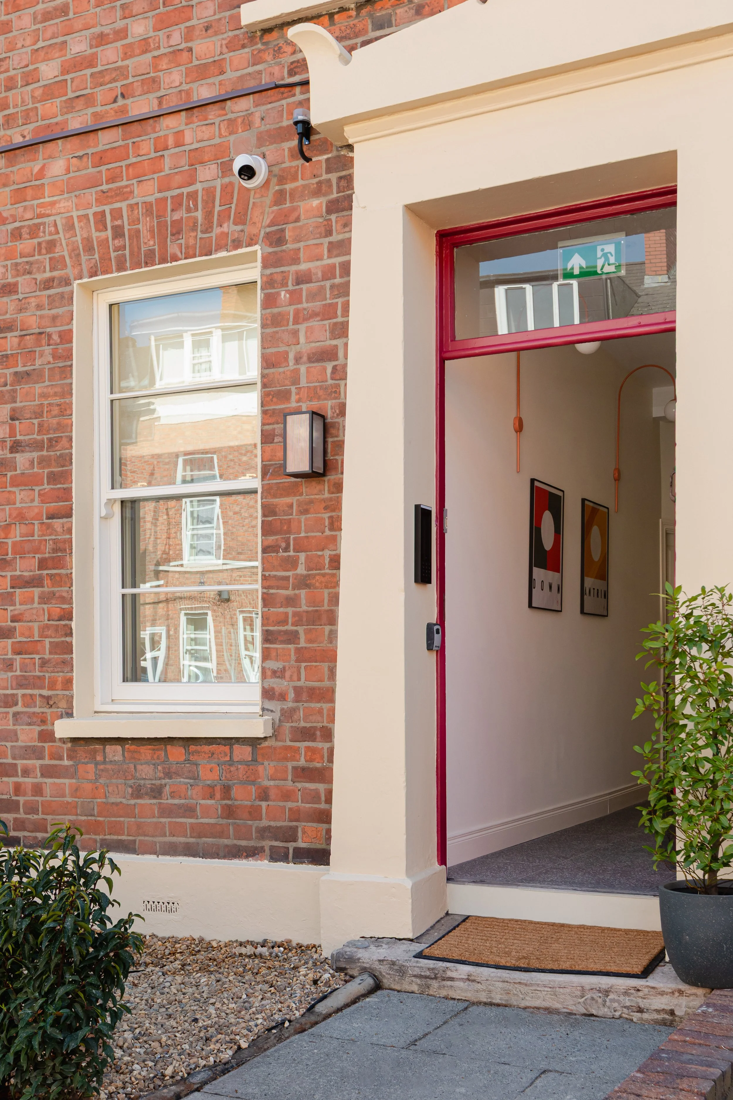 View of an apartment entrance with a red doorframe and an indoor space visible through a glass panel. The entrance is decorated with two framed abstract art pieces on the wall, a potted plant, and wall-mounted lamps. The entry mat is placed outside on gray paving, with brick and beige exterior walls surrounding the door.