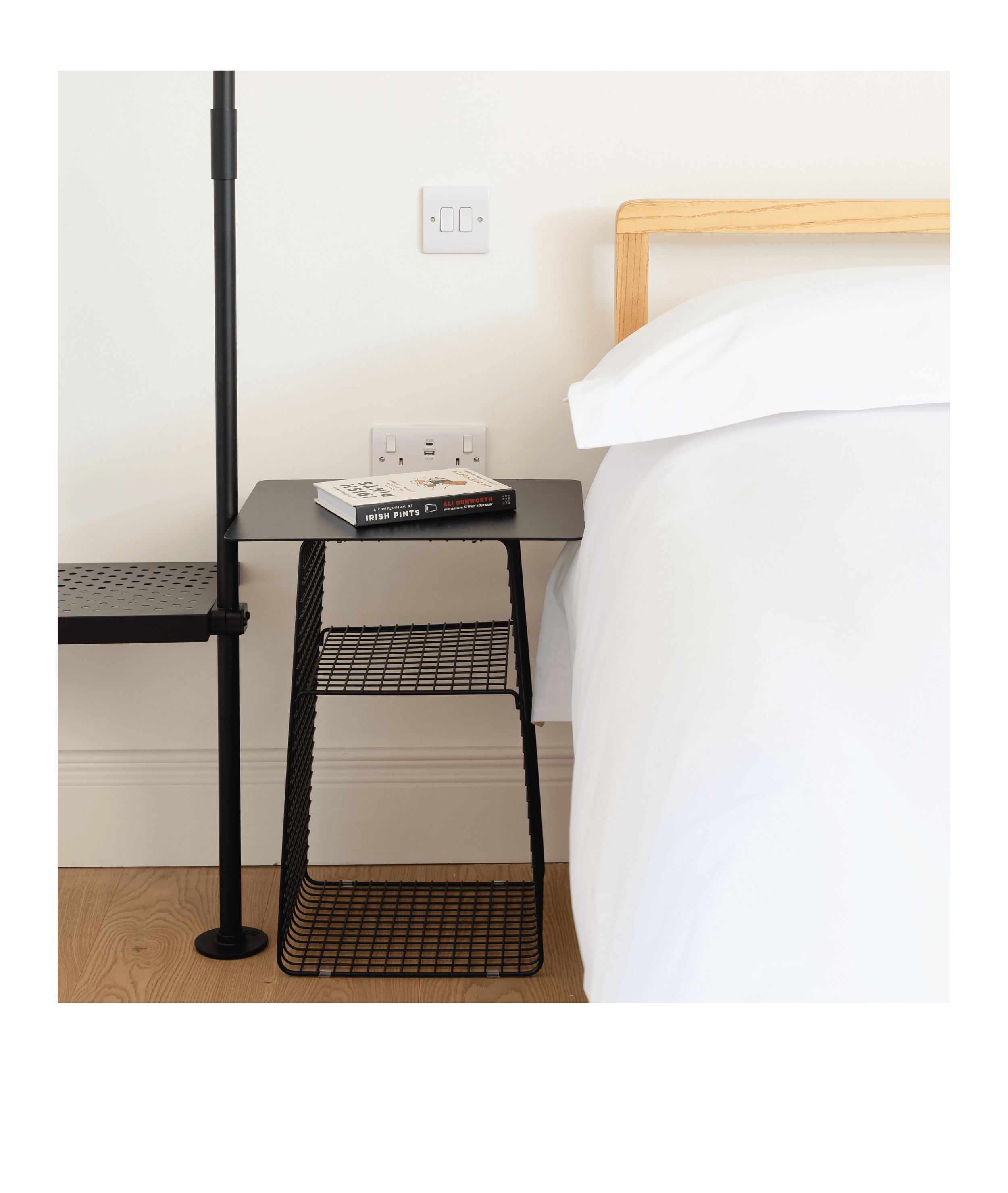 Close-up of a black wire side table with books on top, placed next to a bed with white bedsheets, a wooden headboard, and a wall outlet with a switch.