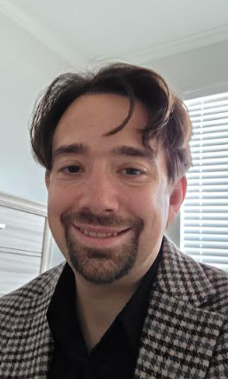 A man with dark brown hair, a goatee, smiling, wearing a checkered blazer and black shirt, in a room with white blinds.