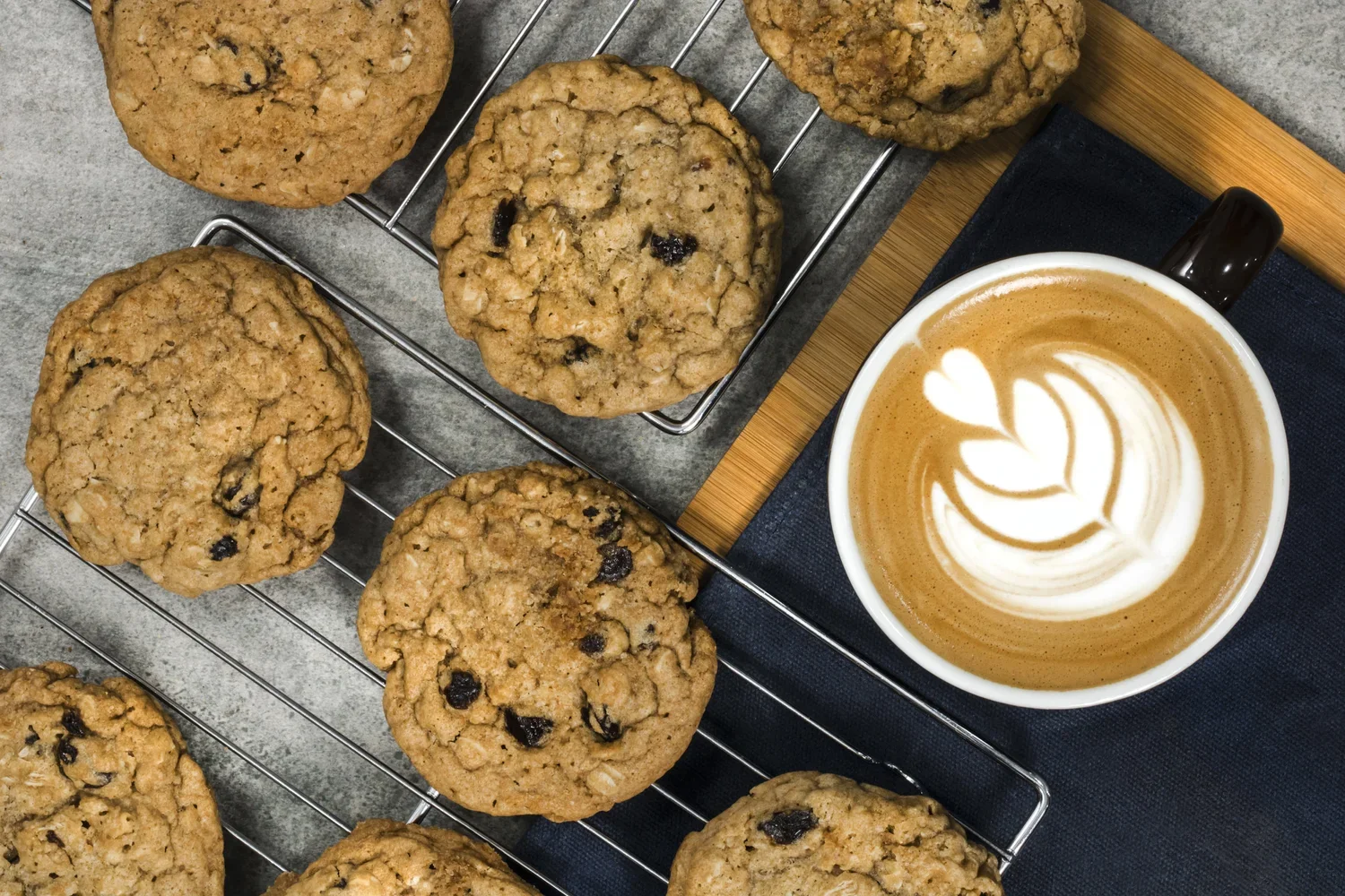 Oatmeal raisin cookies cooling on a wire rack next to a cup of latte with latte art, placed on a dark cloth napkin on a wooden tray.