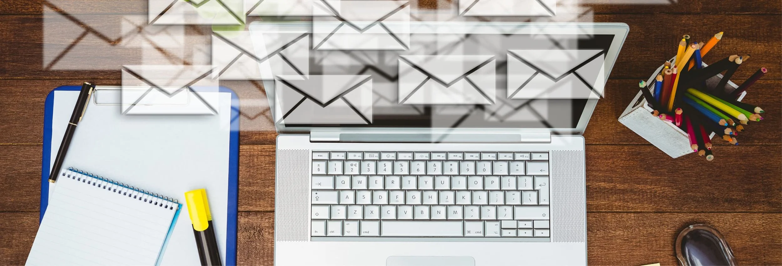 A laptop on a wooden desk with numerous email icons above it, surrounded by a notebook, pen, highlighter, colorful pencils in a holder, and a coffee mug.
