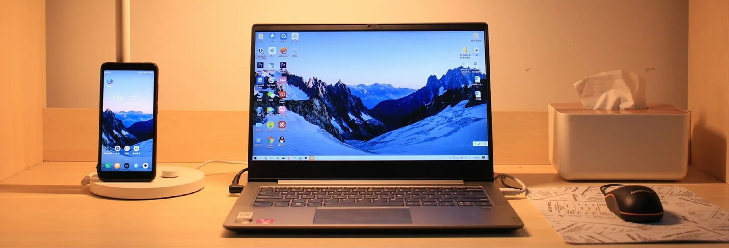 A workplace desk with a smartphone on a wireless charging pad, a laptop with a mountain landscape wallpaper on the screen, a computer mouse on a piece of paper, and a tissue box in the background.