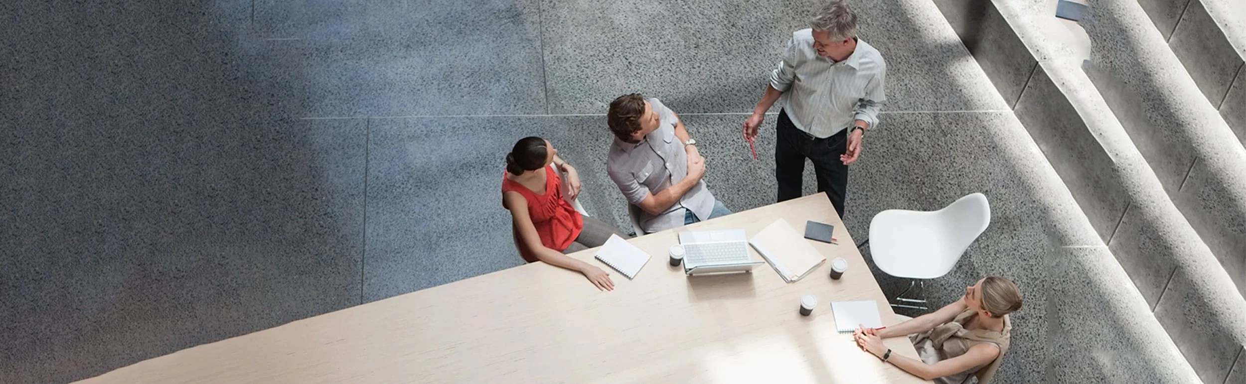 A top-down view of four people standing and sitting around a conference table in a modern office space, engaged in conversation.