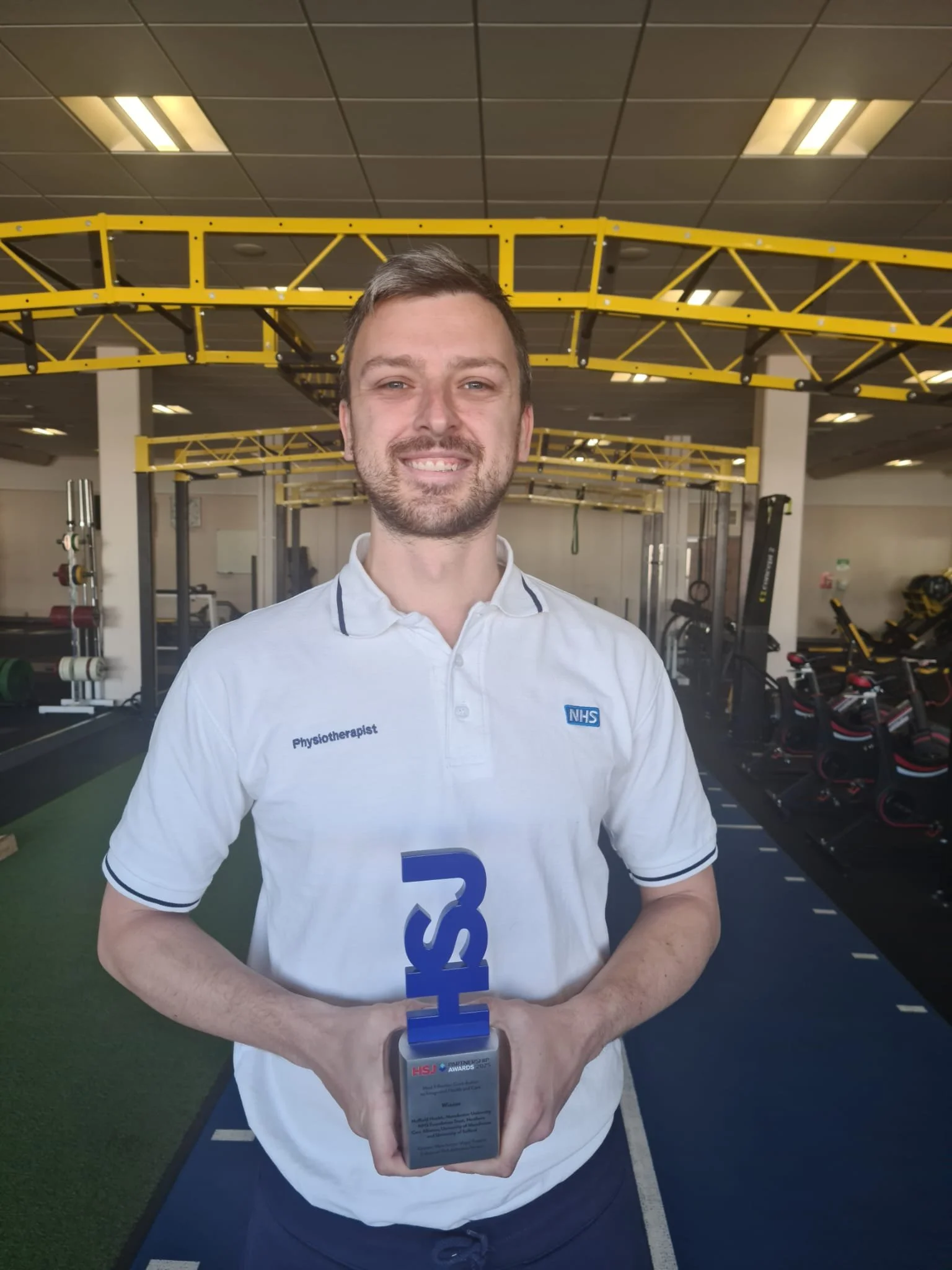 A man in a white NHS physiotherapist polo shirt smiling and holding a blue award trophy in a gym.