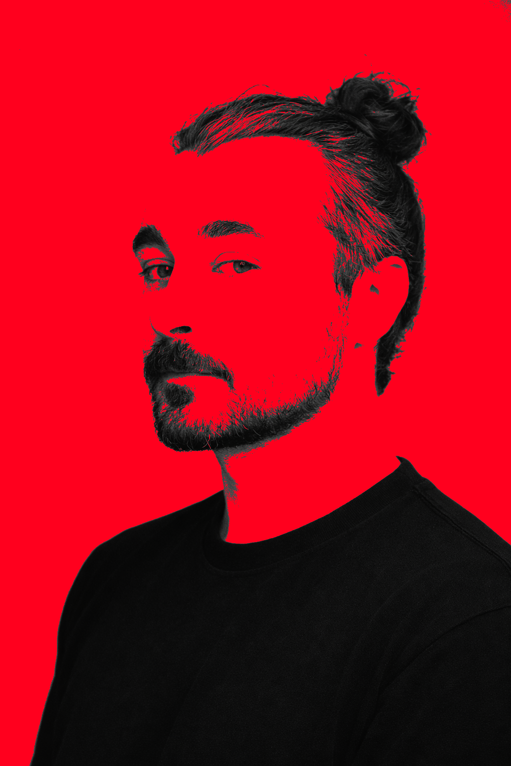 Ritratto di un uomo con barba, capelli raccolti in una crocchia, abbigliamento nero, sfondo rosso