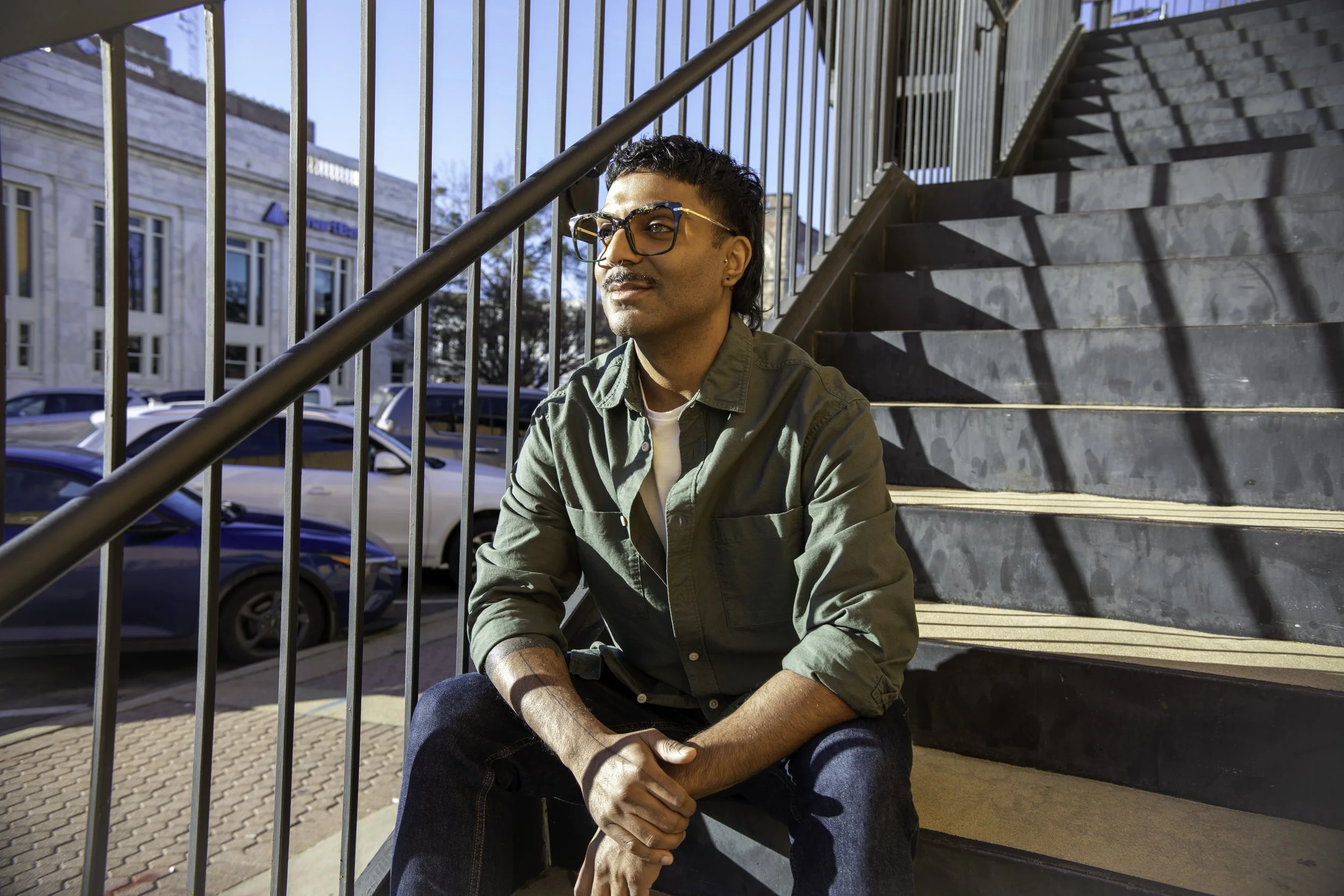 A man sitting on outdoor stairs with metal railings, wearing sunglasses and a green jacket, looking off into the distance, with a building and parked cars in the background.