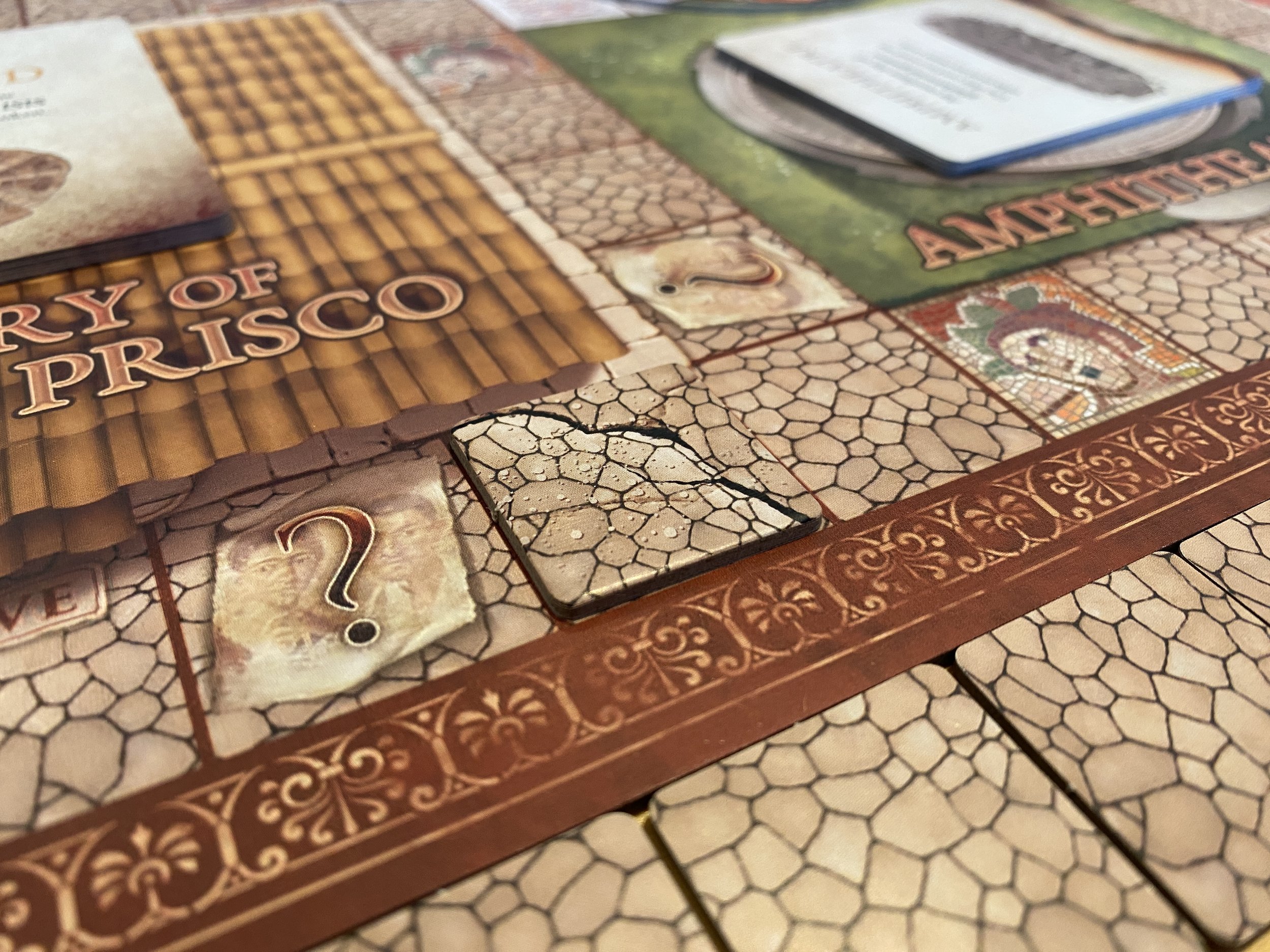A photo of the Escape From Pompeii board game showing a game board with tiles beside it. One tile has been placed onto a square of the board, making the space look like it has been cracked in half