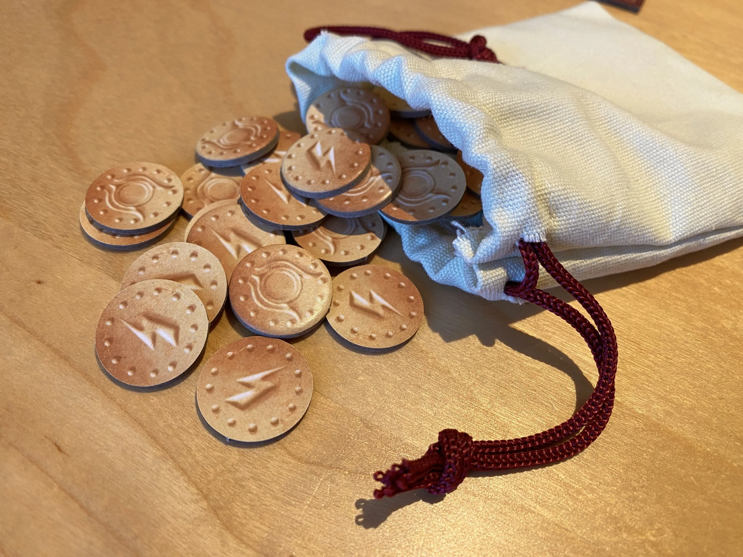 Gold tokens from the Escape From Pompeii board game, with two symbols on, spill out of a cotton bag with dark red drawstrings