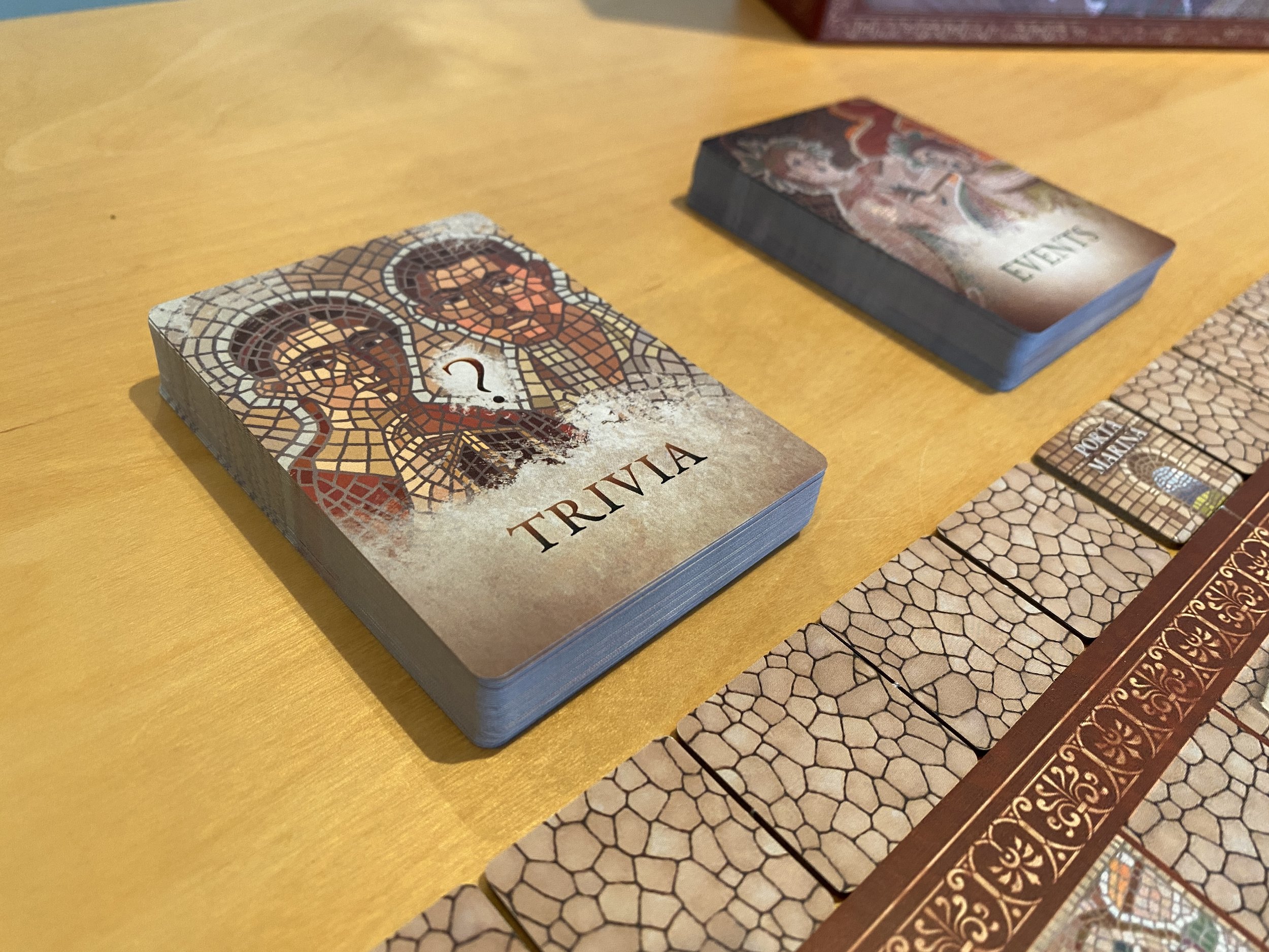 Two decks of cards with mosaic artwork sit beside the game board of Escape From Pompeii. One reads 'trivia', the other reads 'events'