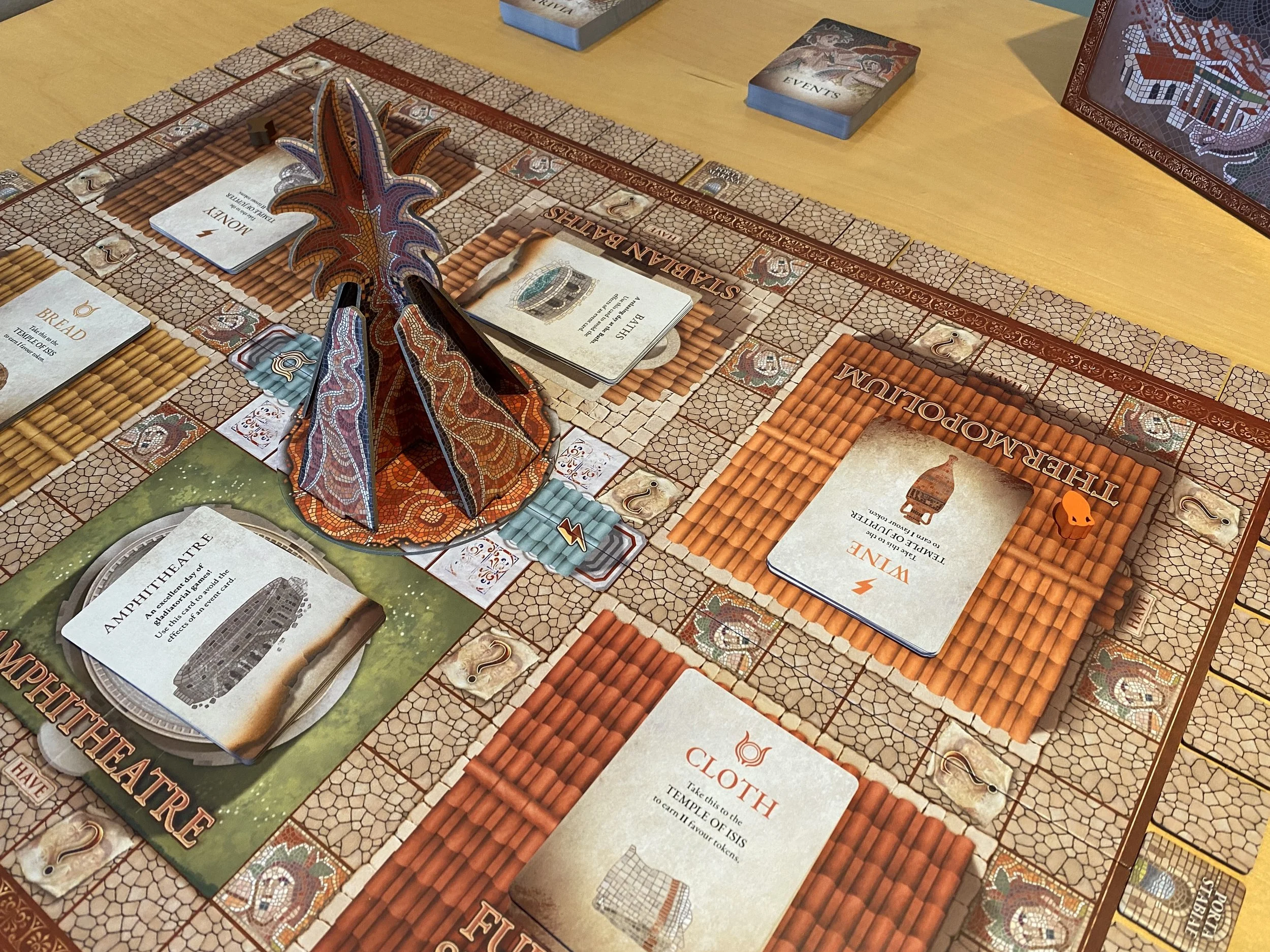 A board game with cards, wooden playing pieces, and a 3D cardboard volcano on. There are tiles around the edge of the board and two decks of cards at the side. The artwork has a mosaic style