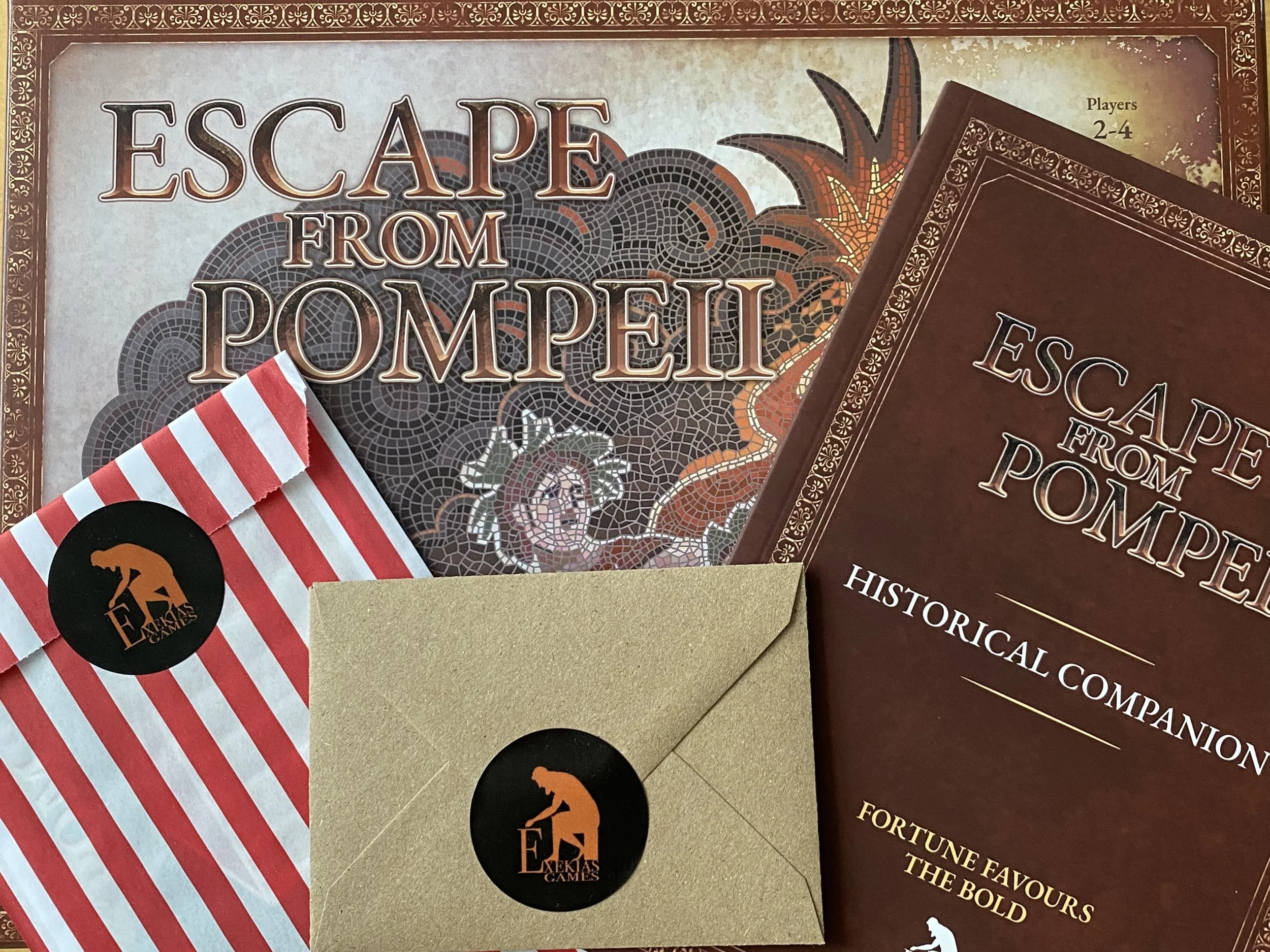 A game box with mosaic artwork and text reading 'Escape From Pompeii'. On top are three items: a book reading 'historical companion', a red and white paper bag sealed with an Exekias Games sticker, and a small brown envelope