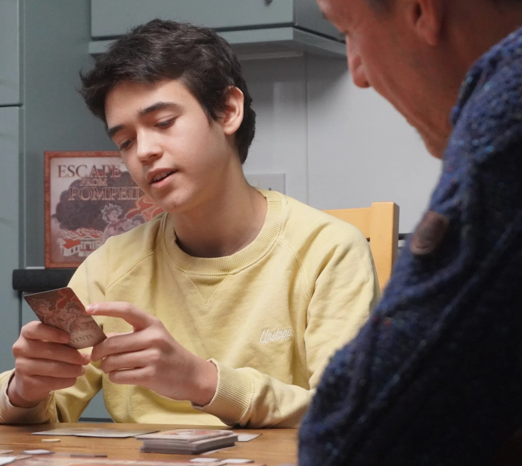 A child reading a card. The back of the card has a mosaic design and reads 'events'. On the table in front on them is a board game. Behind them is a box reading 'Escape From Pompeii'. An adult is sitting to the right