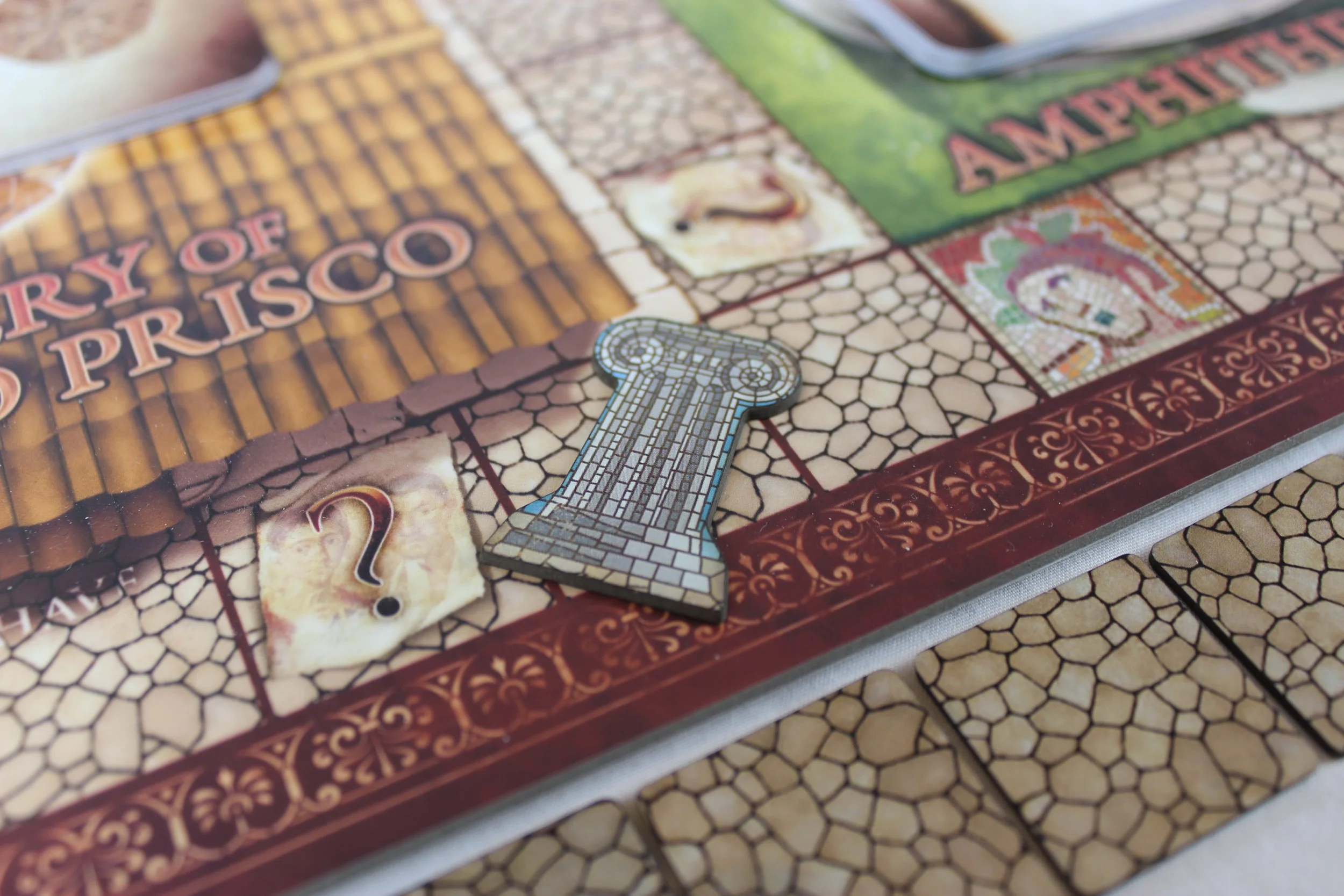 A photo of the Escape From Pompeii board game showing a board with tiles beside it. A token shaped like an ionic column in a mosaic style is placed on one of the squares
