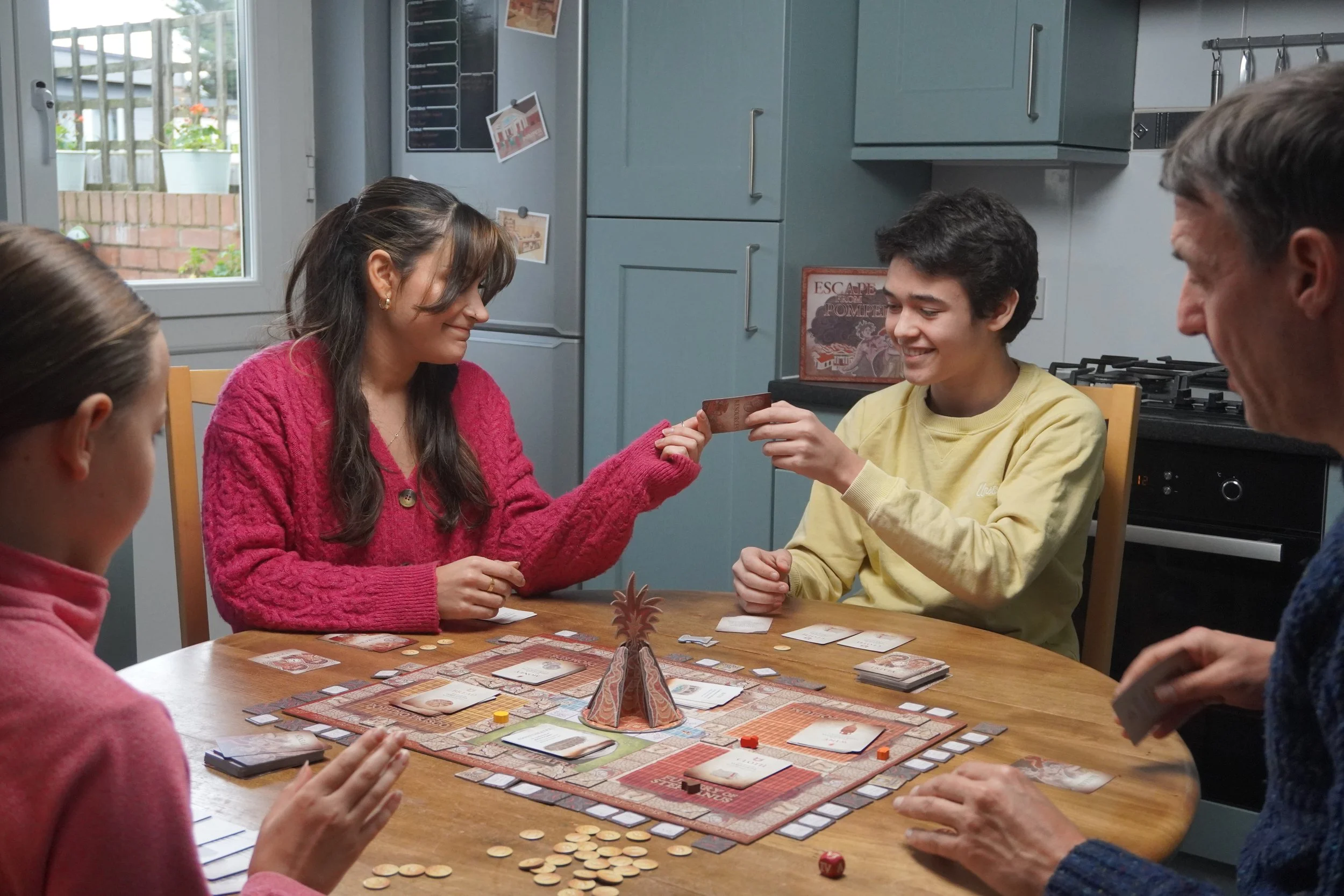 A family playing a board game. An adult hands a child a card. The game box in the background reads Escape From Pompeii
