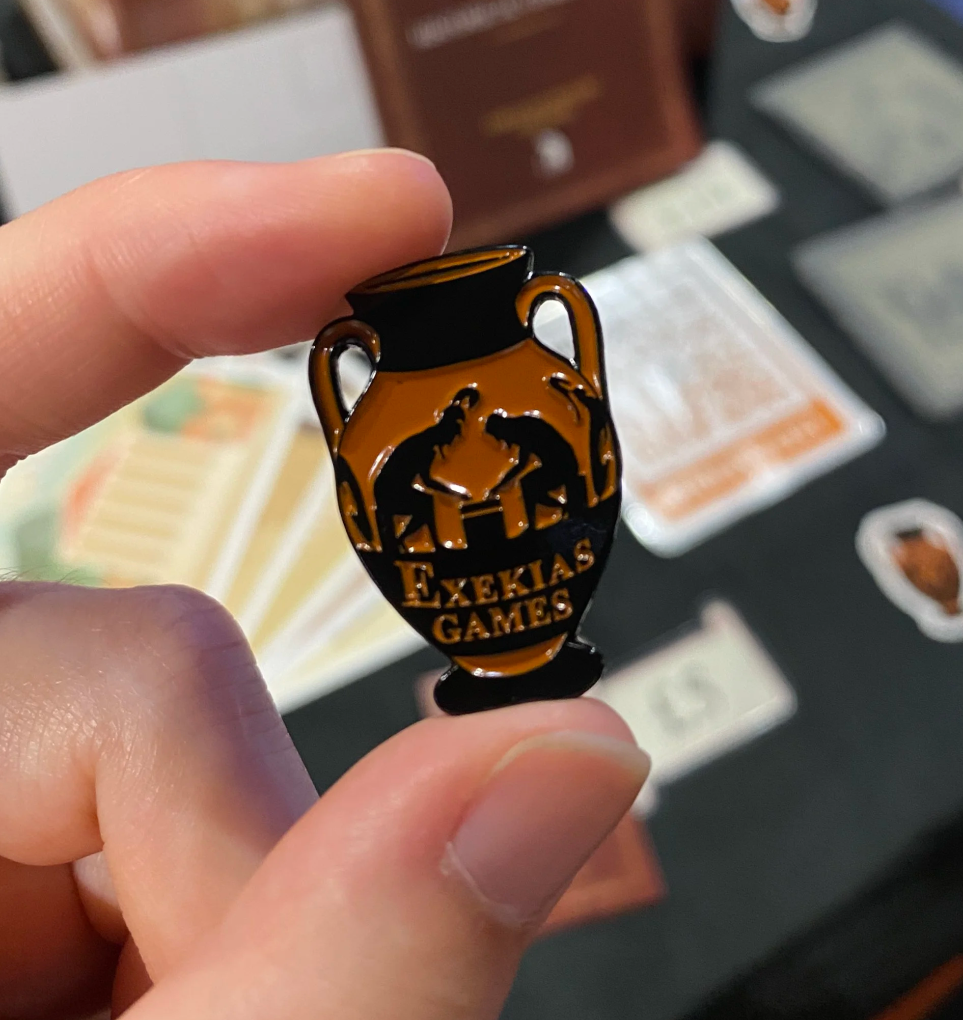 A pin badge in the shape of a Greek vase, with the words 'Exekias Games'. The design on the vase is styled after the ancient Greek vase of Ajax and Achilles playing a board game by the painter Exekias