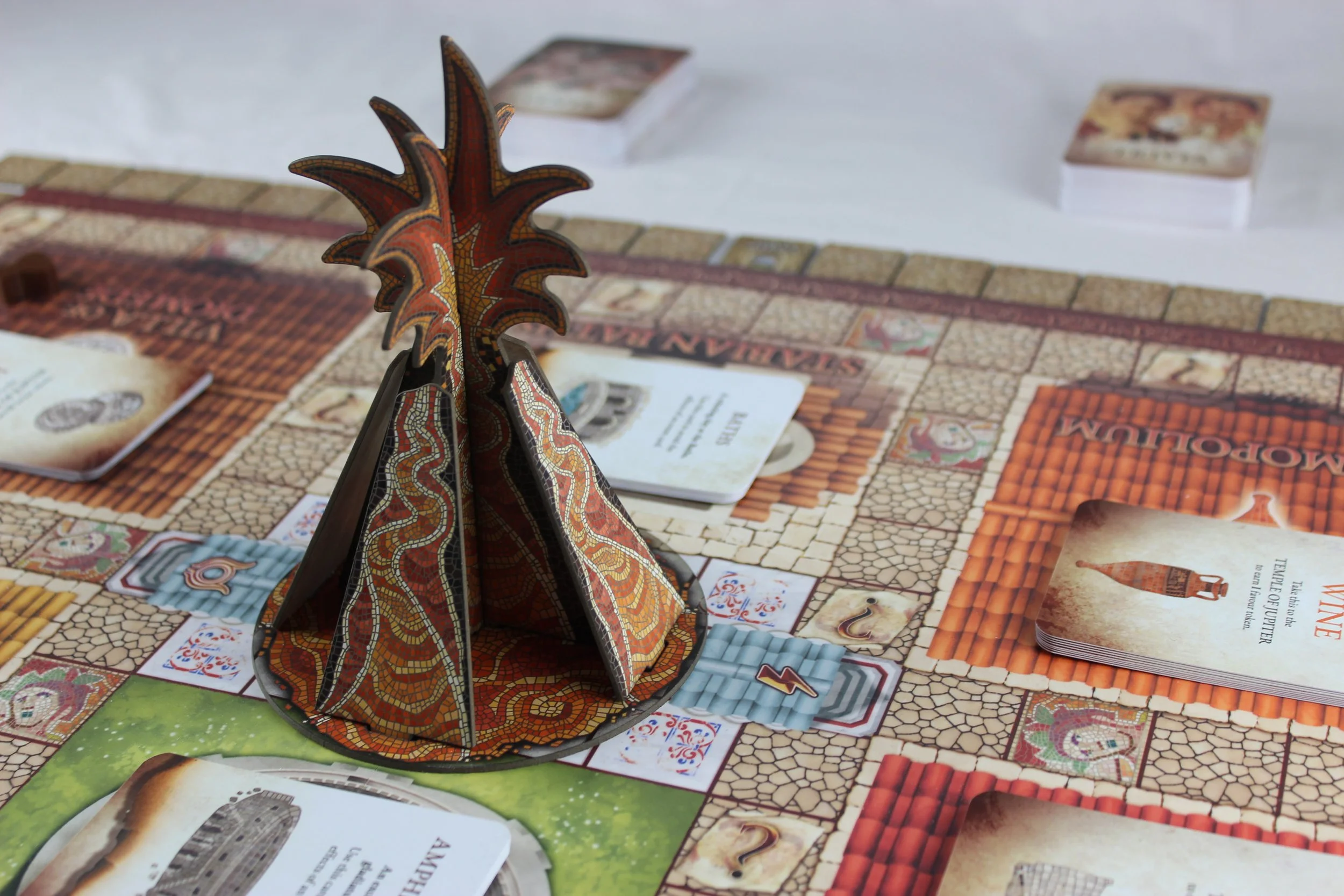 The board of the Escape From Pompeii board game, with a 3D cardboard volcano with mosaic artwork in the centre, and cards around the board