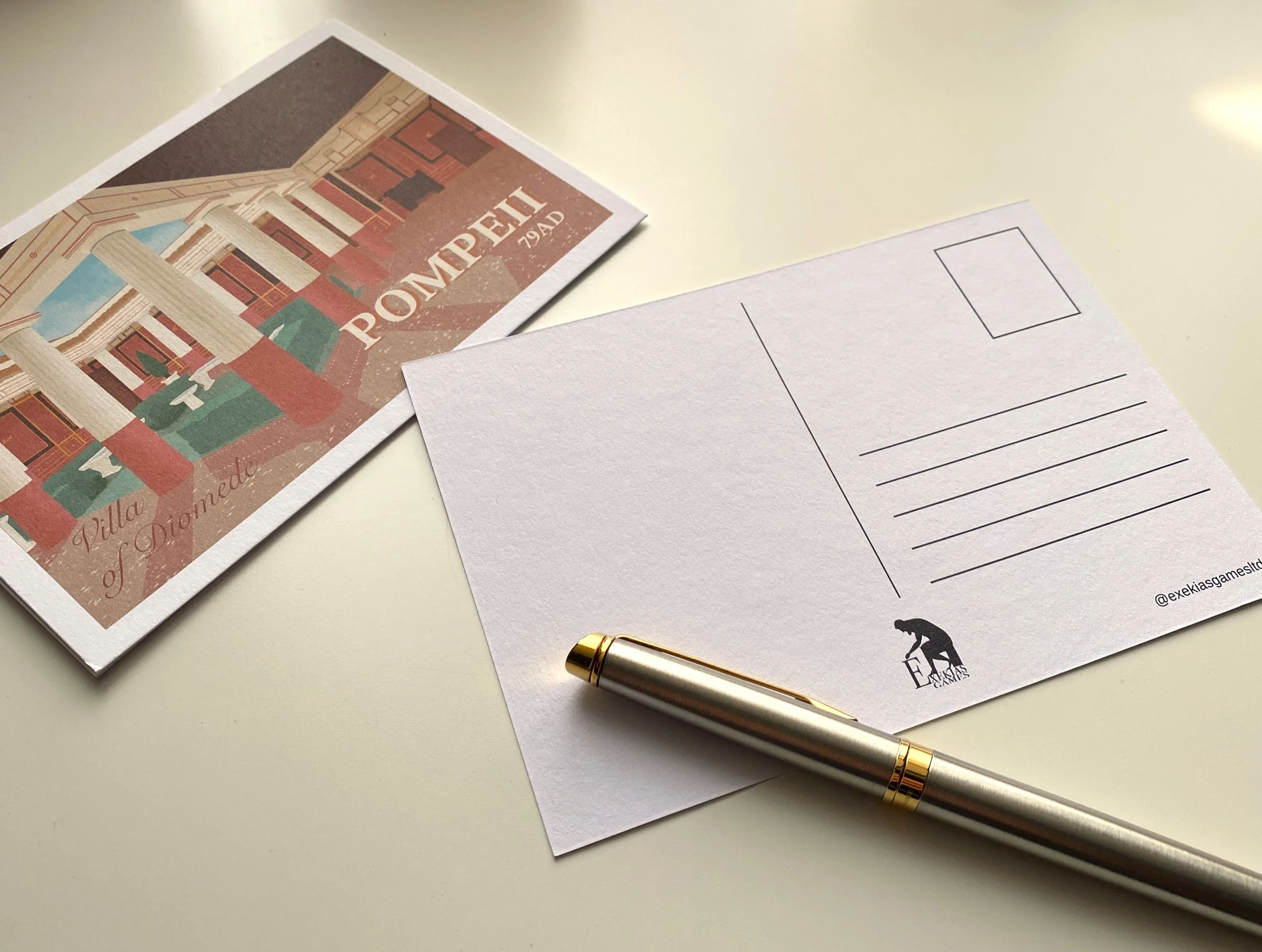 A 30s-style postcard of a reconstruction of the Villa of Diomede in Pompeii on a desk. Beside it is the back of another postcard and a fountain pen