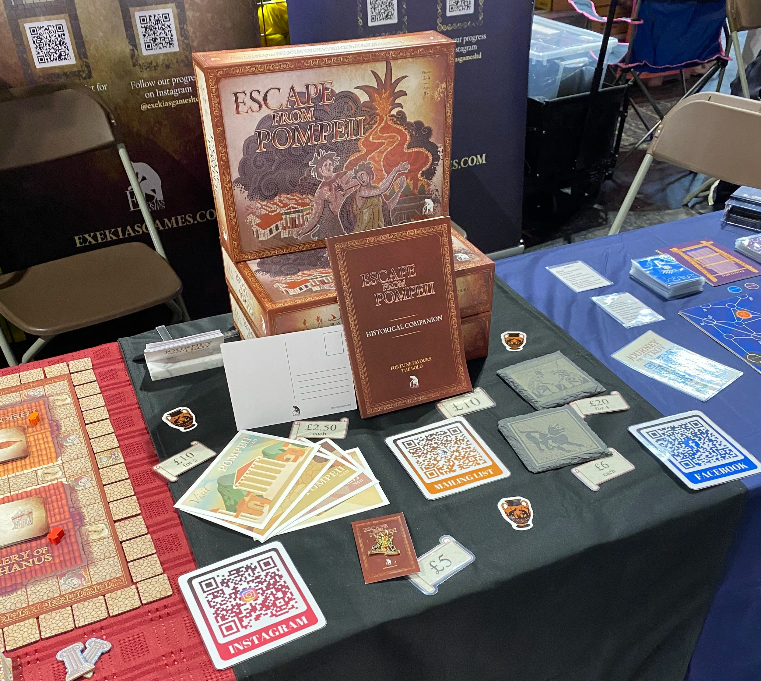 A table displaying the Escape From Pompeii board game as well as several extras, including postcards, a book, coasters, and a pin badge. To the sides are games laid out to demonstrate