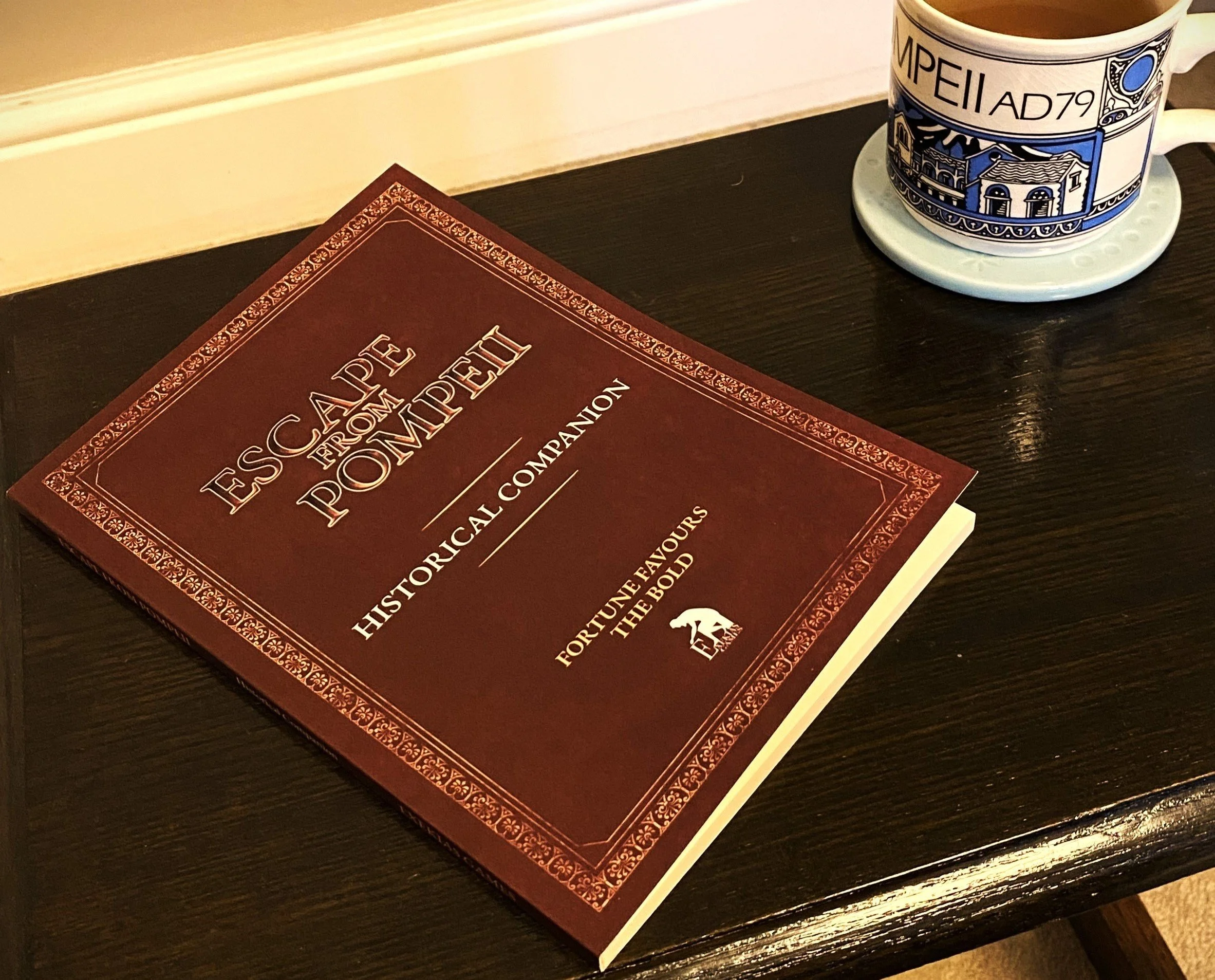 A book on a coffee table reading 'Escape From Pompeii: historical companion. Fortune favours the bold', with an Exekias Games logo. There is a cup of tea on a coaster next to it