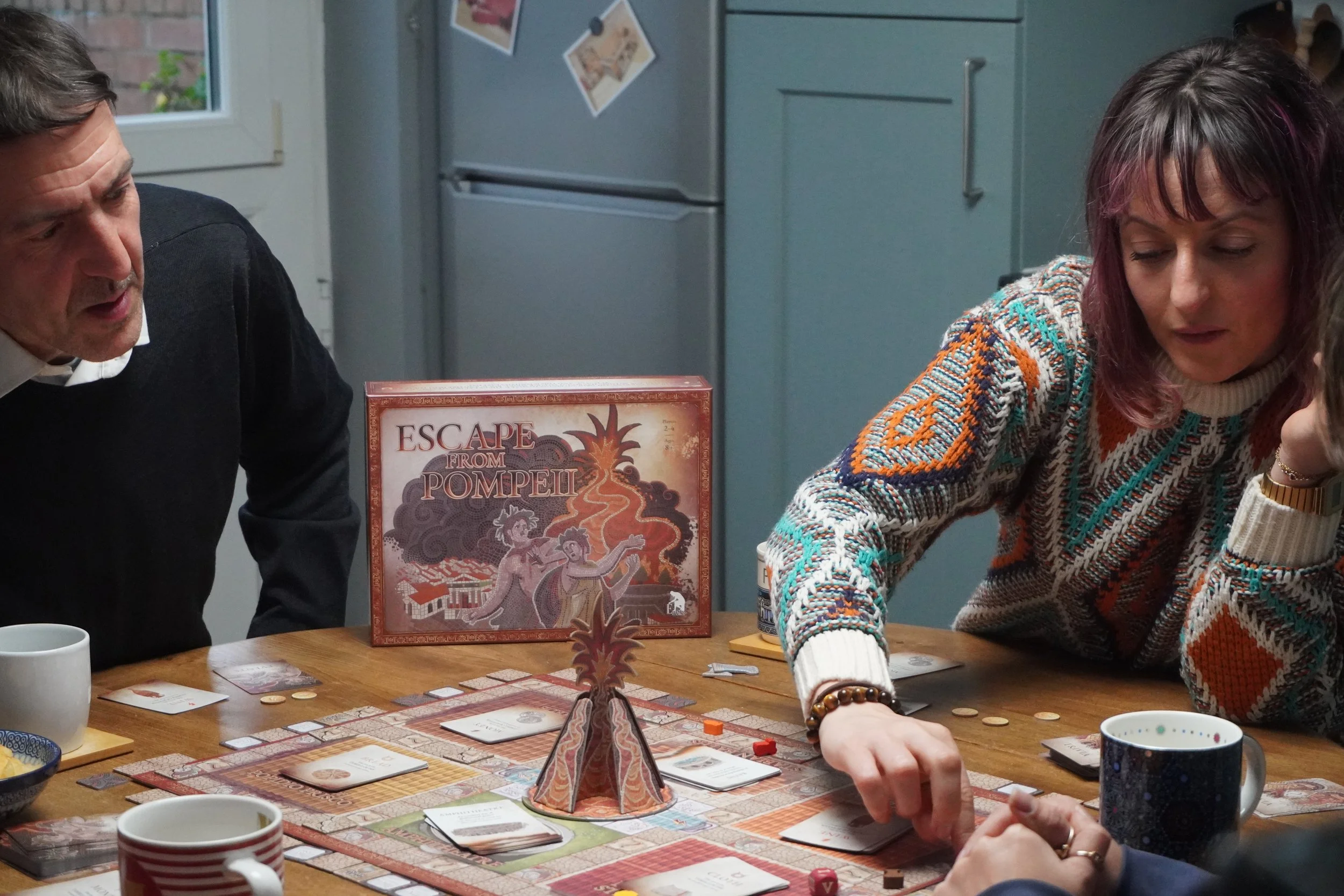 Four adults playing a board game with mugs and snaacks on the table. The game box reads Escape From Pompeii