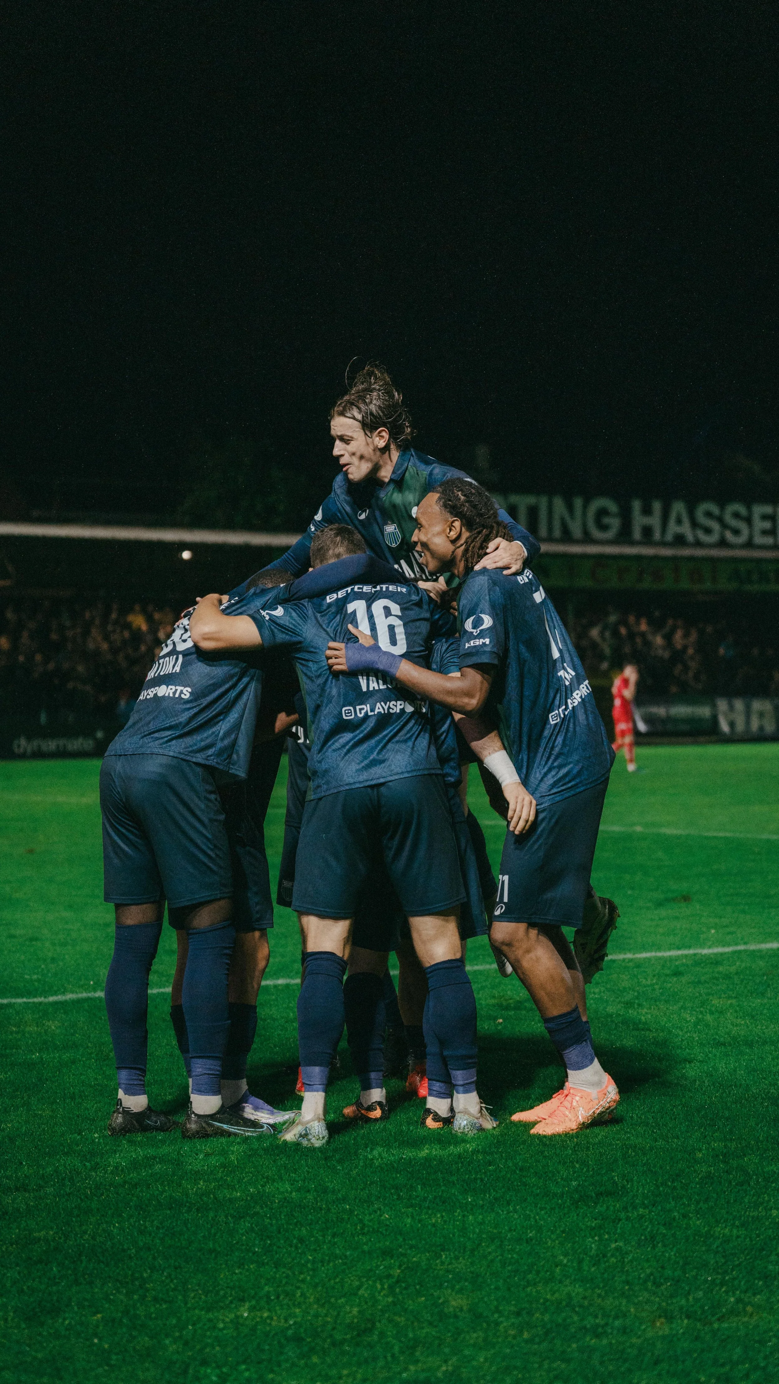 Soccer players in blue jerseys celebrate during a match, with one player being lifted by teammates on a green grass field at night.
