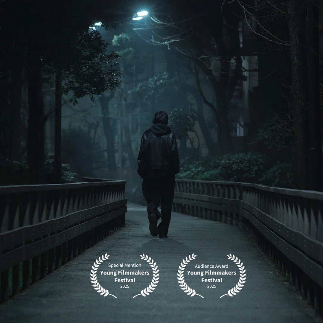 A person walking alone on a dimly lit wooden bridge at night, surrounded by trees in a park or forest, with award laurels at the bottom for the Young Filmmakers Festival 2025.