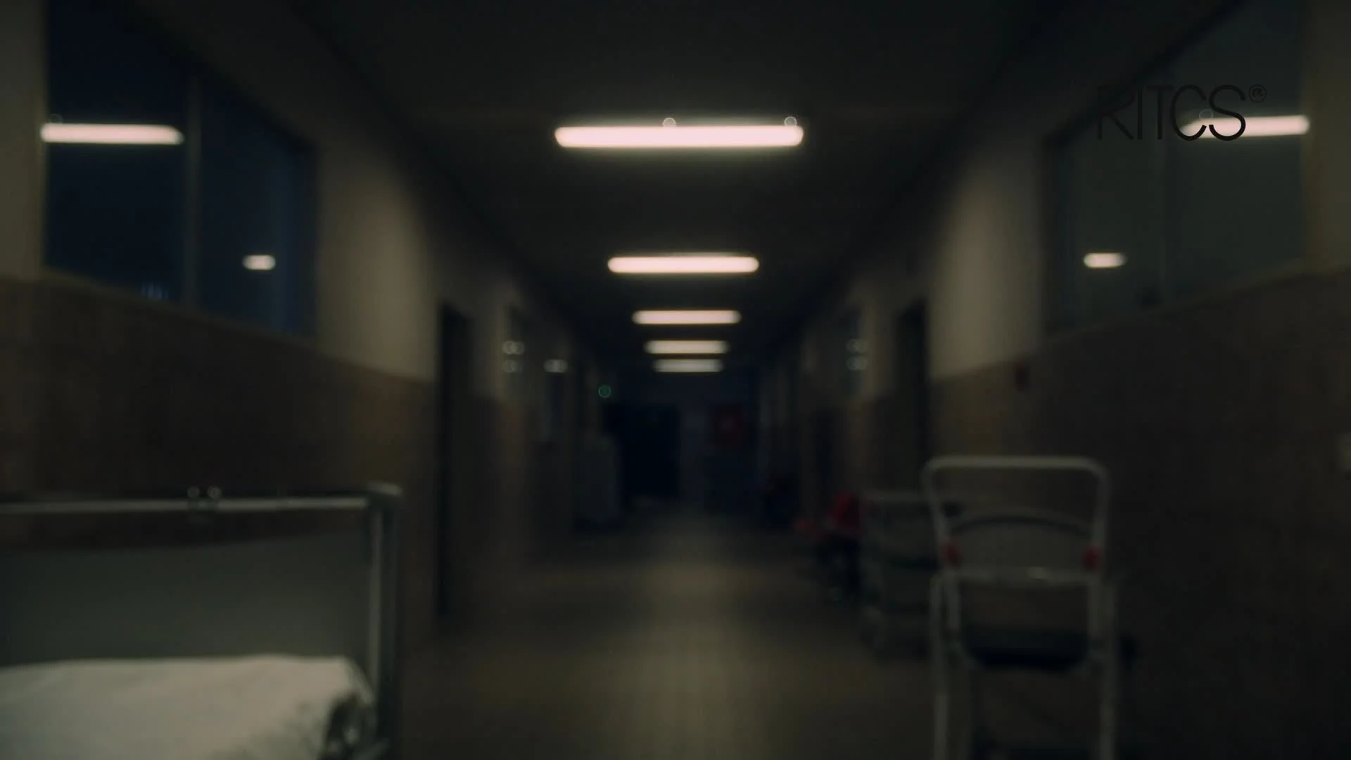 Dimly lit hospital corridor with hospital beds and medical carts on the side.