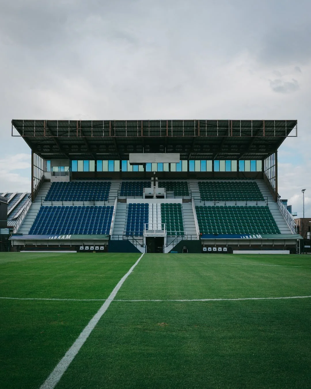 Empty soccer stadium with green field and blue and green seating, under cloudy sky.