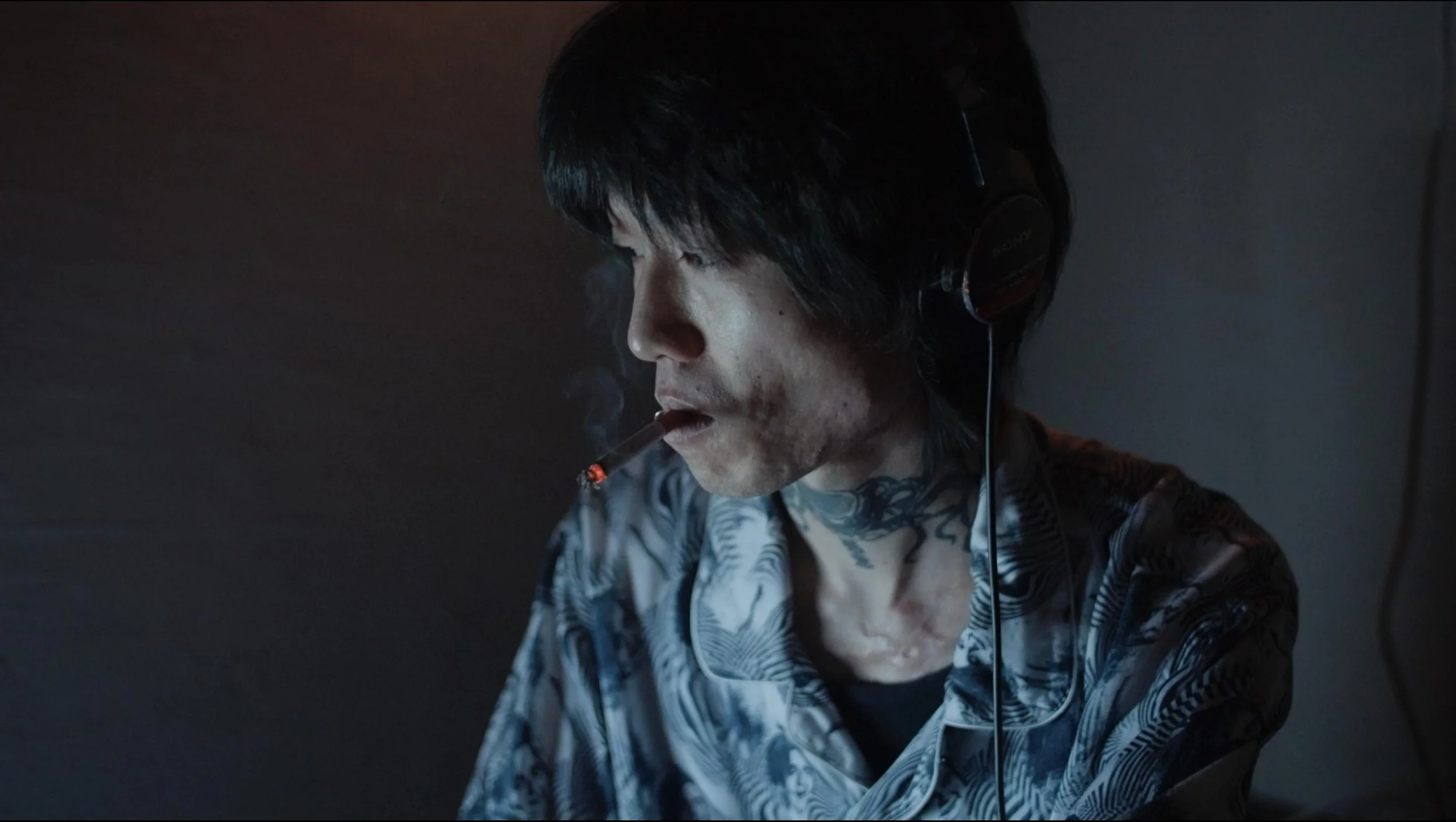 A person with black hair wearing headphones and a patterned shirt, smoking a cigarette in a dimly lit room.