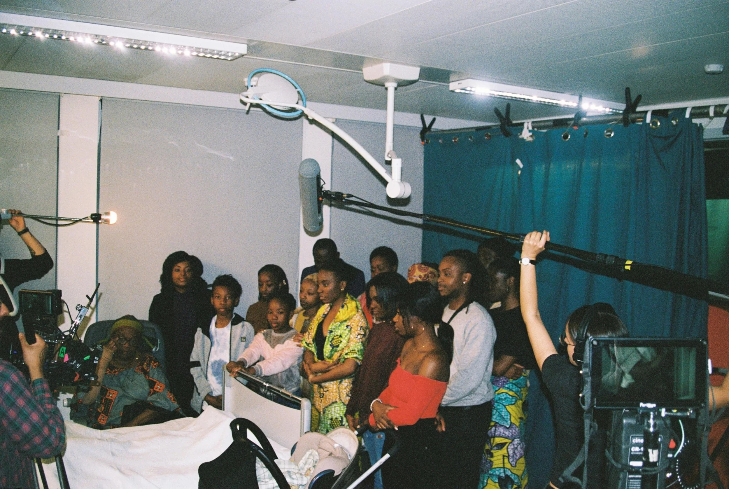 Filmmaking crew filming a scene in a hospital, with actors and a baby in a bed, filming equipment, and a crew member holding a boom microphone.