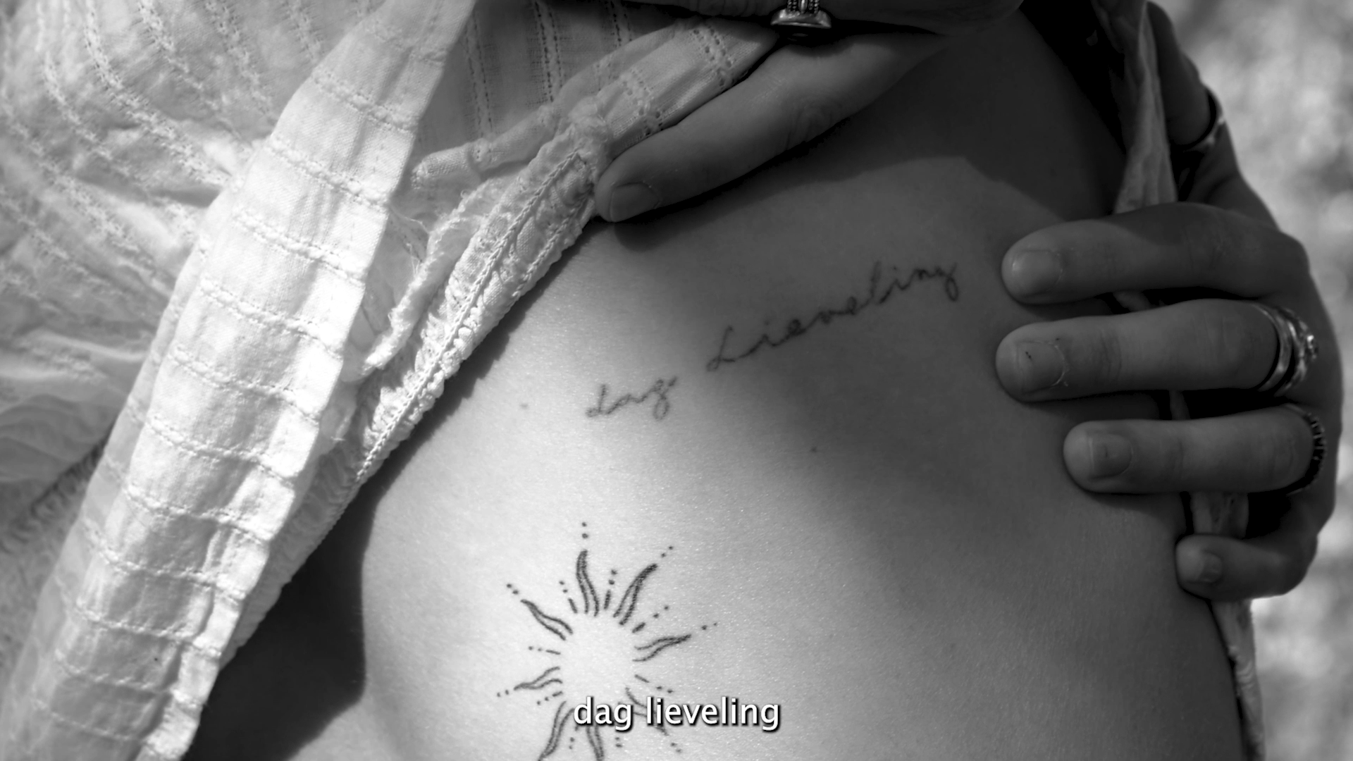 Close-up of a person's side with two tattoos: one is a handwritten phrase 'dog lievling' above a sun design, and the other is a cursive phrase 'lay llieving' on the person's ribcage. The person is wearing rings and a textured, white, open shirt.