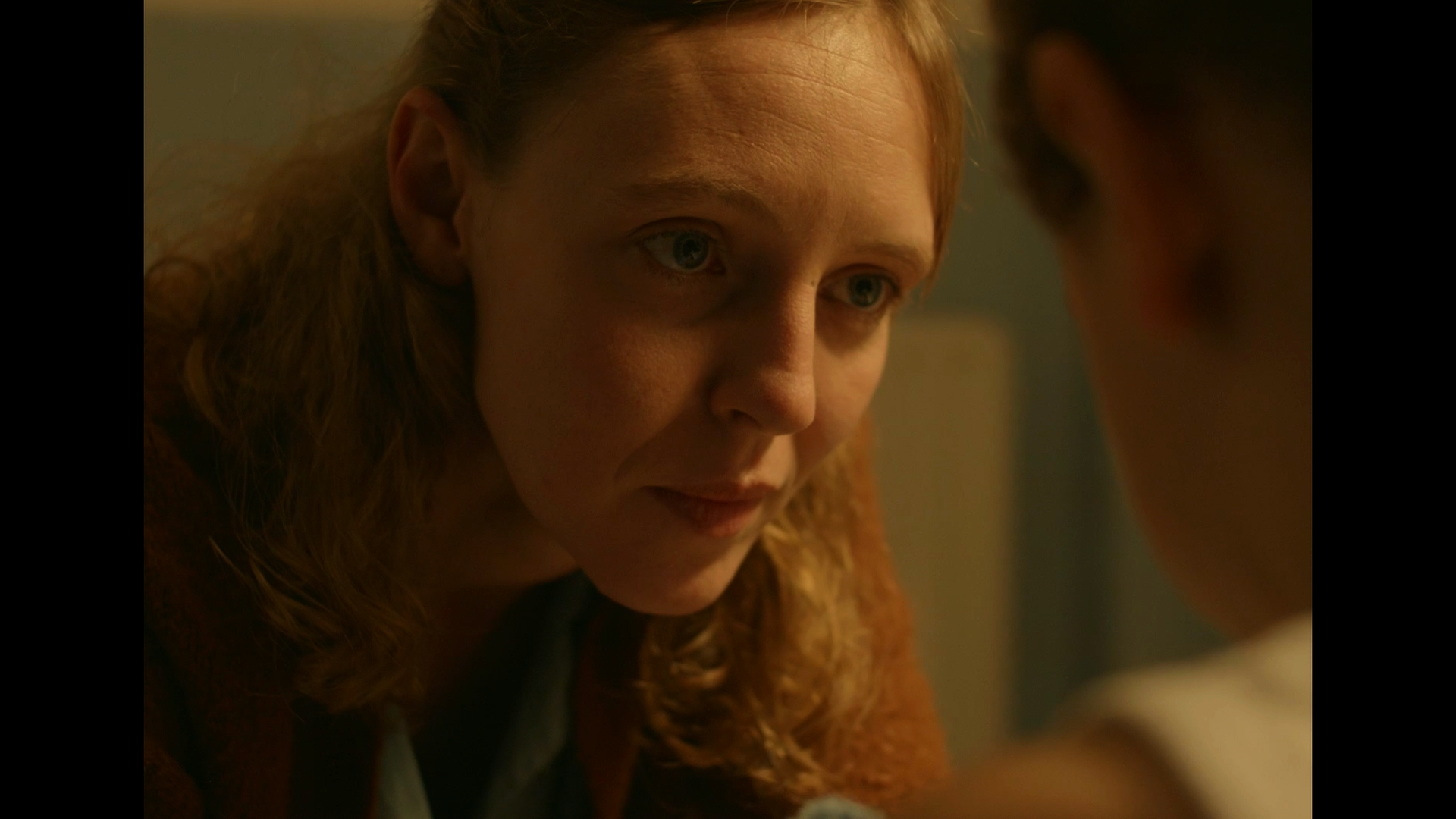 A close-up of a woman with red hair and fair skin looking intently at someone or something outside the frame, with a serious expression.
