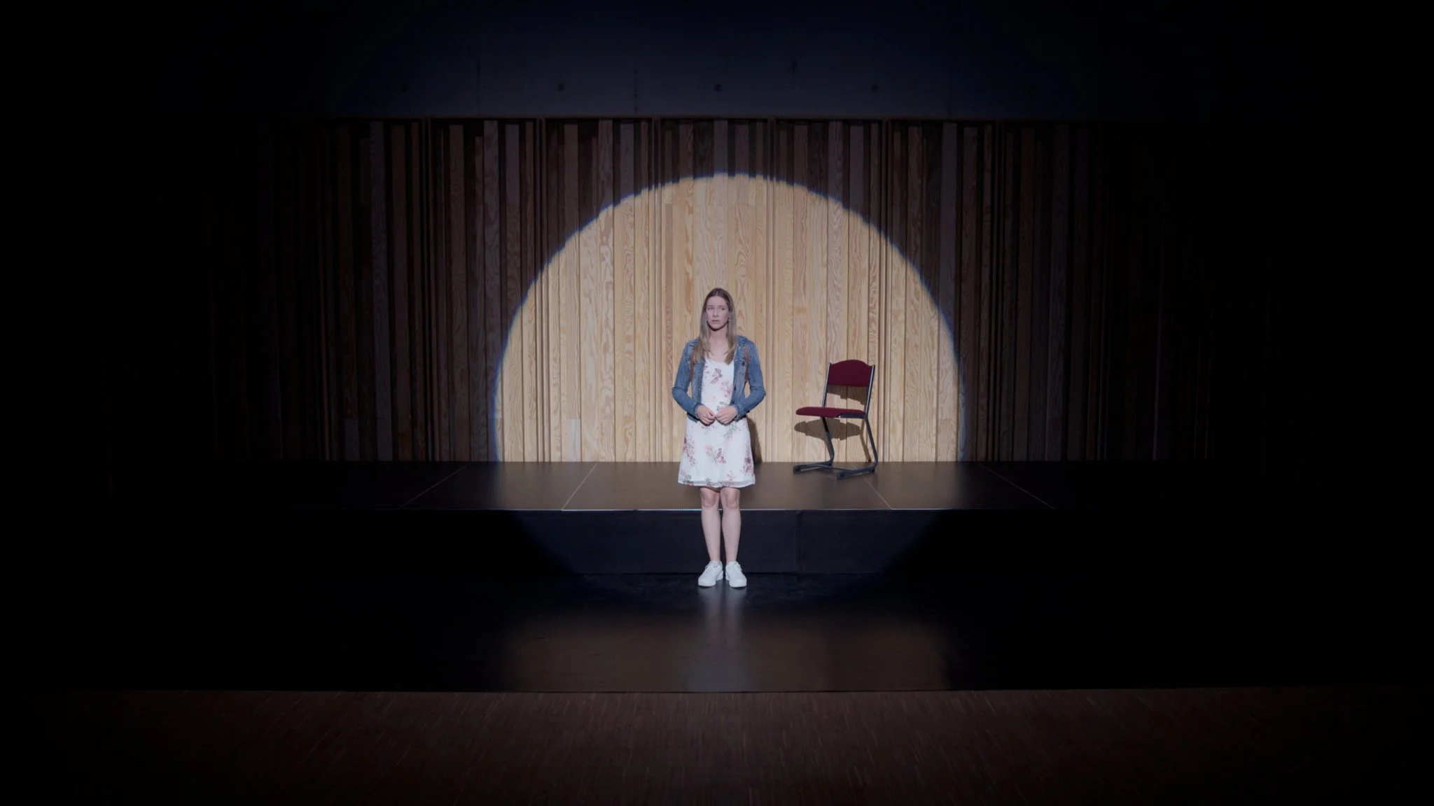 A young woman stands alone on a small stage with a wood-paneled backdrop, illuminated by a spotlight; a red chair is positioned to her right.