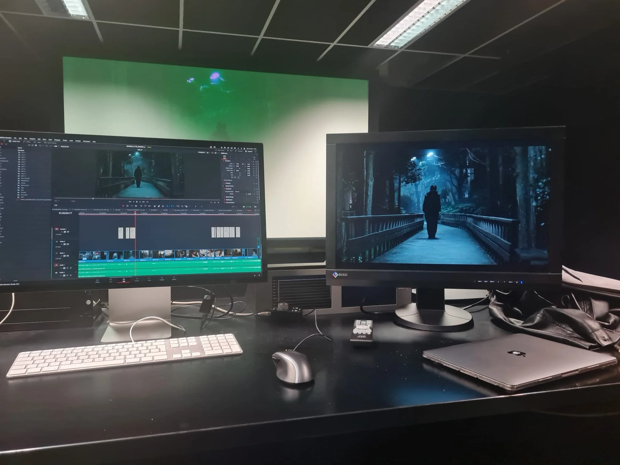 A computer workstation with dual monitors, a wireless keyboard, and mouse. The left monitor displays an editing software with a video timeline, and the right monitor shows a scene of a person walking on a bridge in a forest at night. There is a closed laptop and a black bag on the desk.