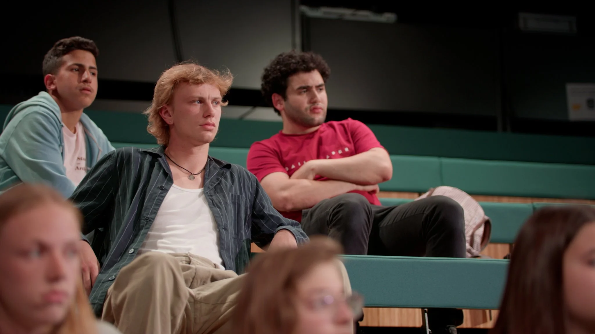 Three teenage boys sitting on theater seats watching an event, with two girls in the foreground.
