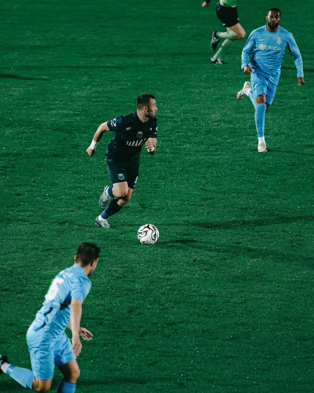 Soccer players on a green field, one in a dark uniform running towards the ball, others in light blue uniforms running after him, during a match at night.
