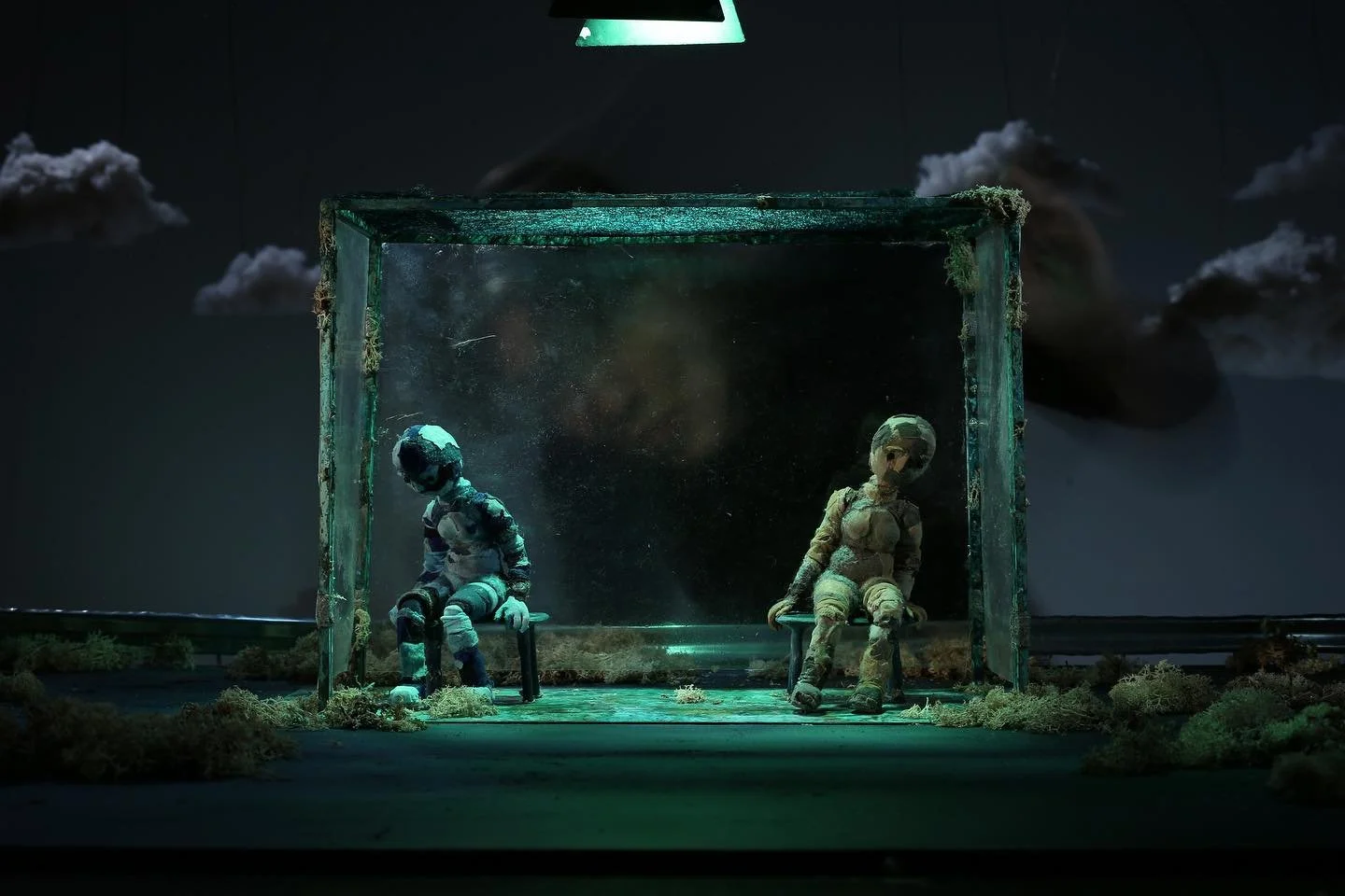 Two astronaut dolls sit inside a glass display case, illuminated by a green light, with a dark, cloudy sky backdrop.