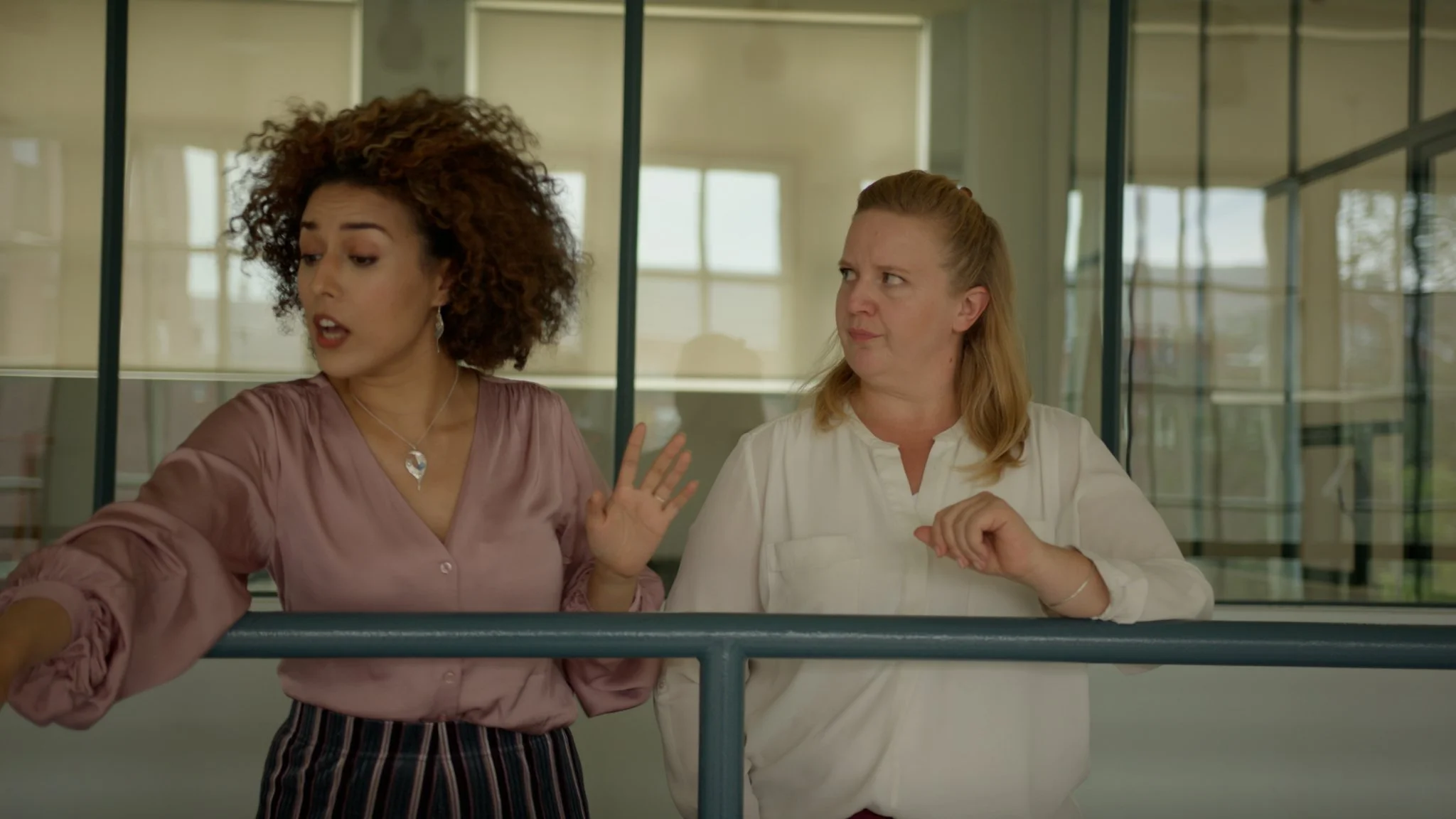 Two women having a conversation indoors, one gesturing with her hand and appearing to speak, the other listening with a skeptical expression.