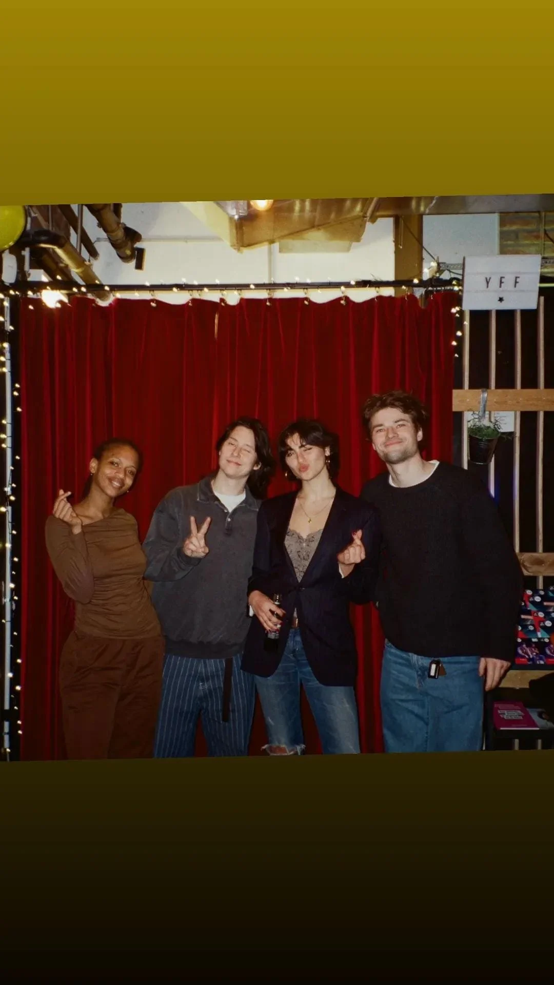 Four friends posing in front of a red curtain with string lights, some making peace signs and finger hearts, celebrating at a social gathering or party.