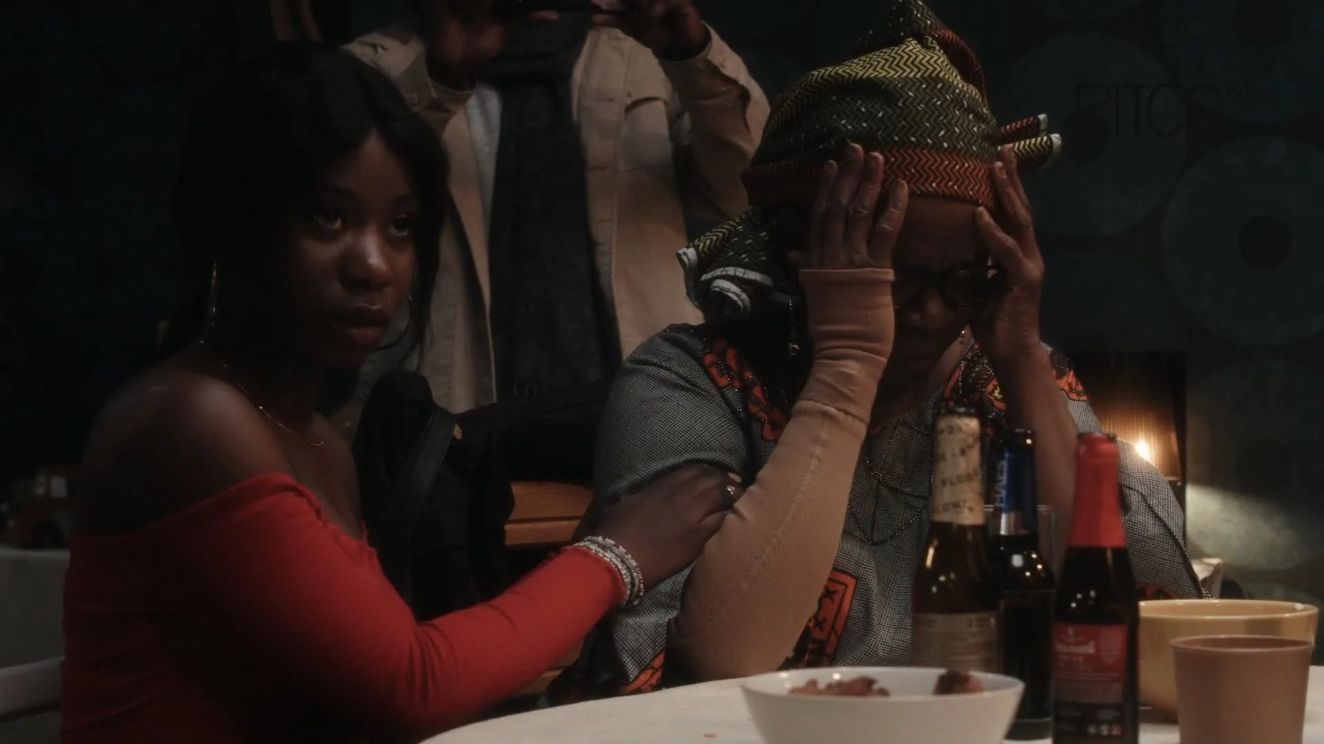 A woman in a red off-the-shoulder top sitting at a table, holding hands with an elderly woman who is covering her face with her hands and wearing a headscarf. There are bottles, bowls, and cups on the table, and a person in the background with a hand on the elderly woman's shoulder.