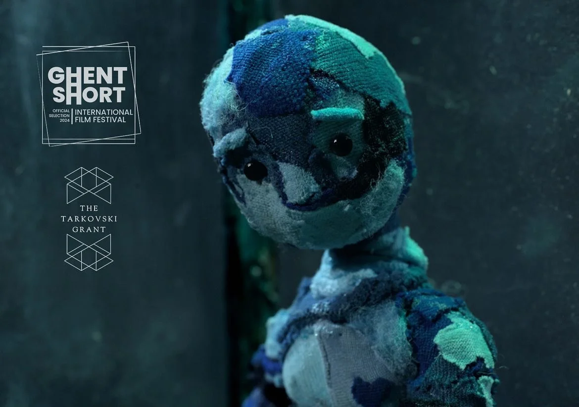 A worn and tattered plush doll with a stitched smiling face, black button eyes, and patchwork clothing, standing against a dark background. The image features logos and text indicating the Ghent Short International Film Festival and The Tarkovski Grant.