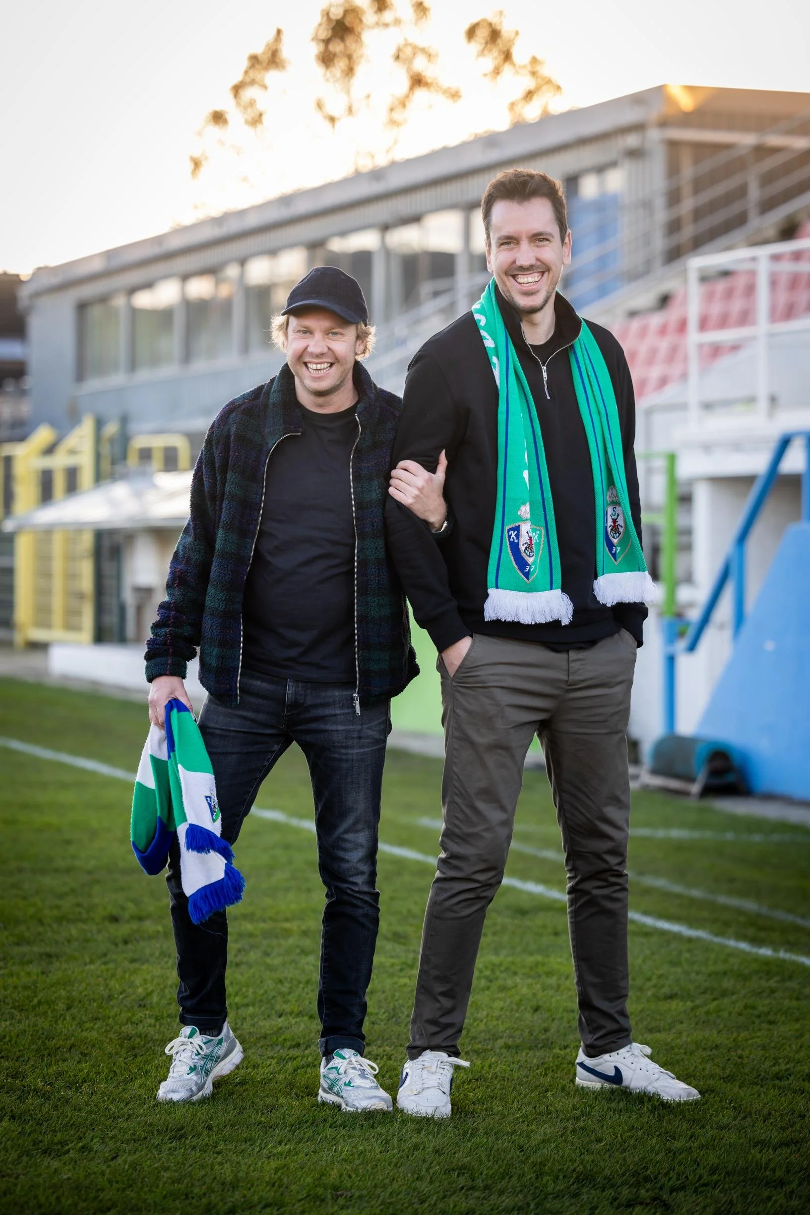 Two smiling men standing on a sports field, one holding a team scarf, celebrating. They are dressed casually, with a stadium and greenery in the background, during sunset.