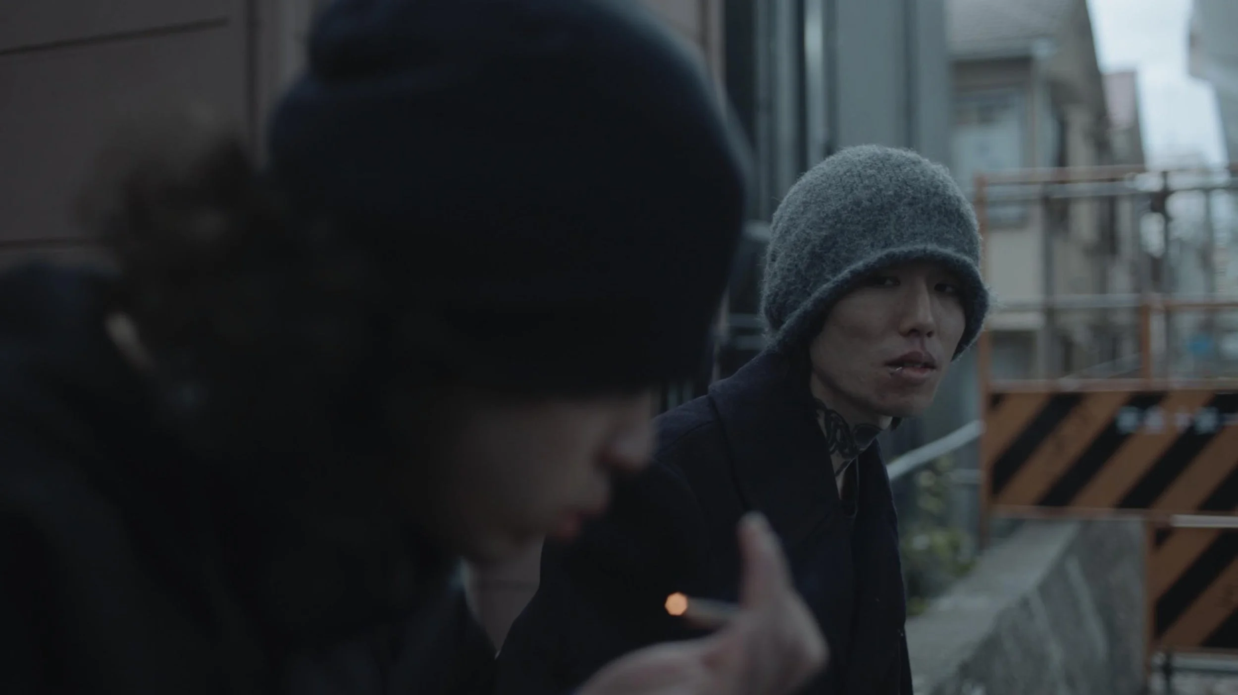 Two individuals sitting outdoors, wearing dark clothing and gray knit hats. The person in the background is looking slightly to the side, with noticeable facial scars and tattoos on their neck, while the other person in the foreground is looking down or away.