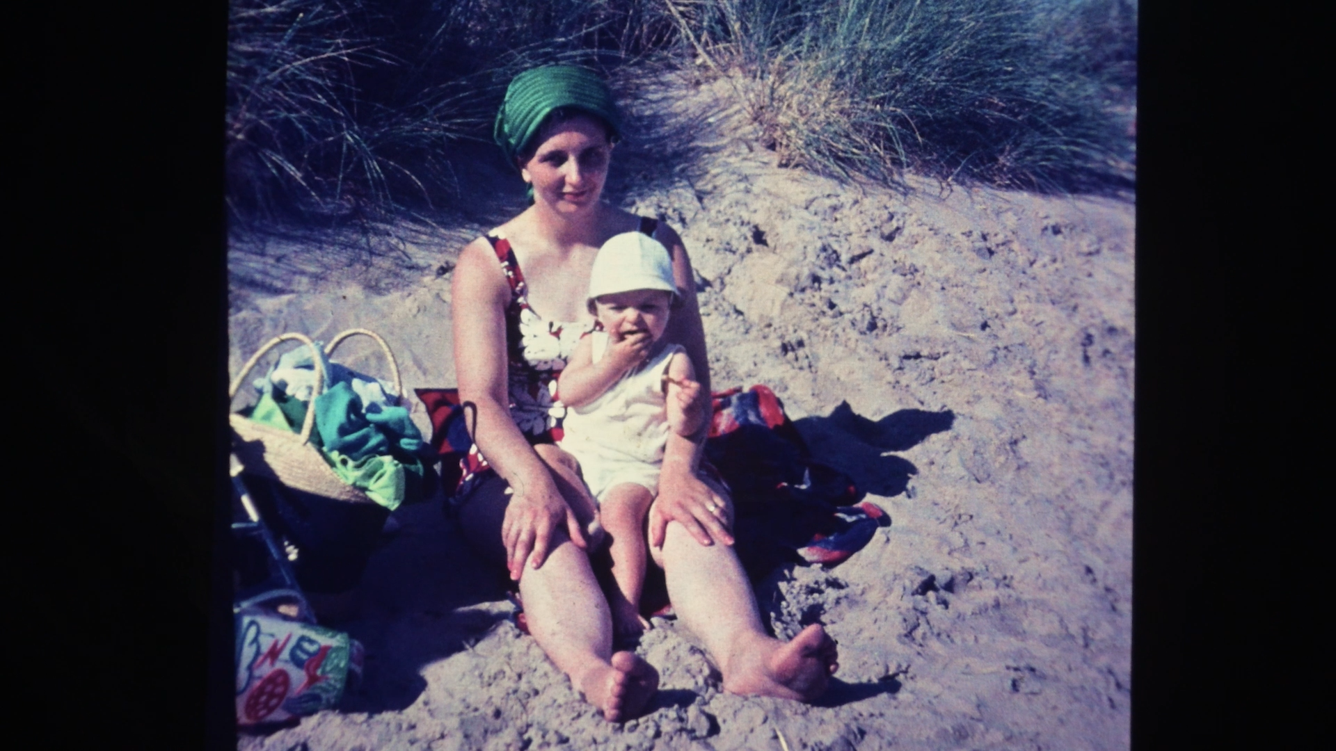 A woman and young child sitting on the sand at the beach, with a basket and bags nearby. The woman is wearing a sleeveless dress with a floral pattern and a green head wrap, while the child is dressed in a white hat and light-colored outfit, with the child biting their finger.