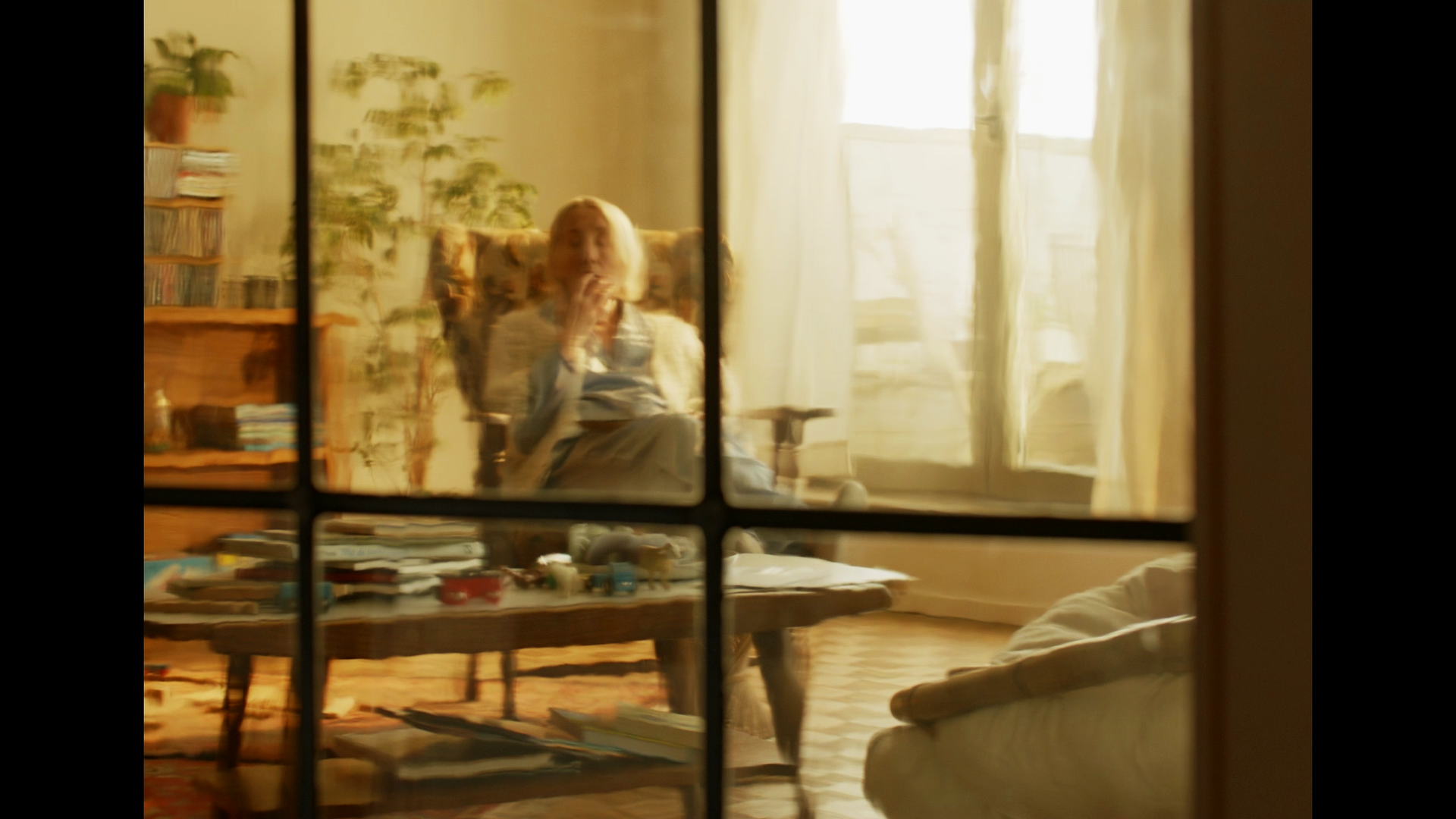 A woman sitting in a chair viewed through a window with four panes, in a cozy room with a bookshelf, a table cluttered with books and toys, illuminated by natural light from a large window with curtains.